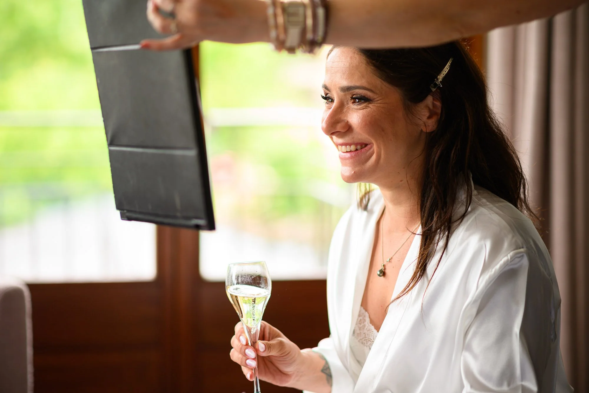 Une femme souriante tient une coupe de champagne dans une pièce lumineuse avec des fenêtres donnant sur un extérieur verdoyant.