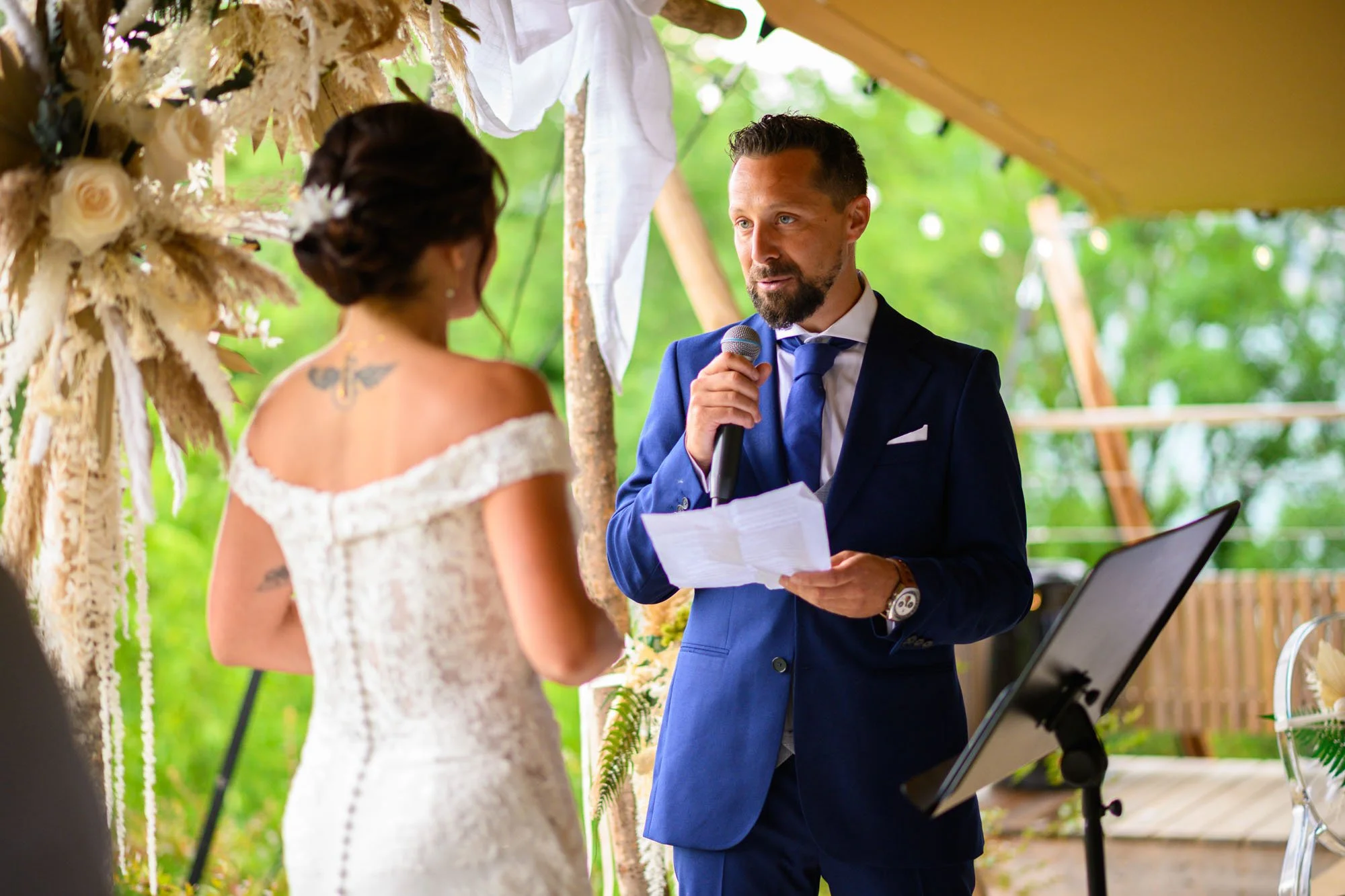 Un homme en costume bleu présente un discours à une mariée lors d'une cérémonie en plein air avec un décor floral.