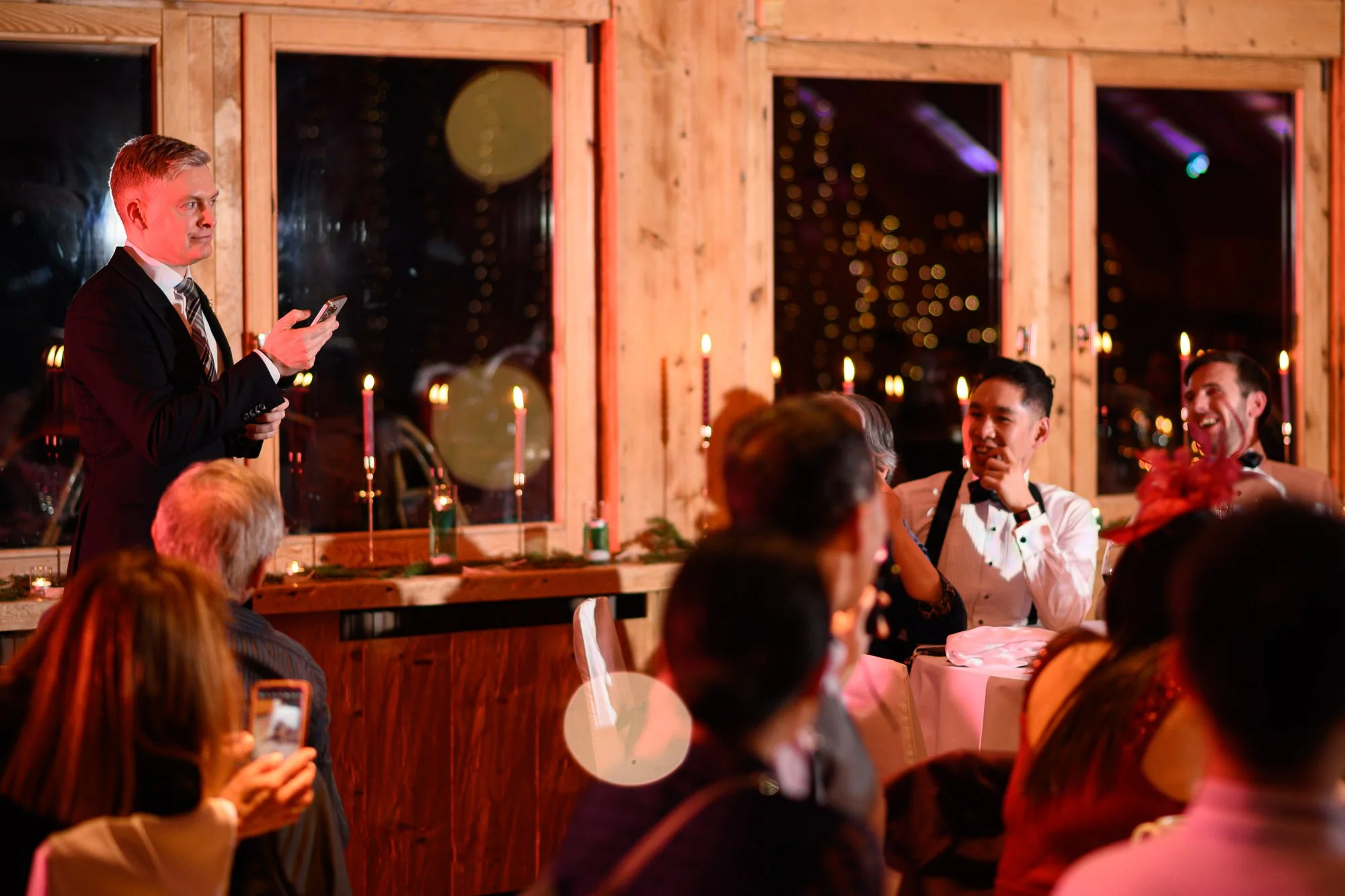 Un homme en costume noir donne un discours lors d'un dîner formel avec des invités assis à une table, dans une ambiance chaleureuse avec bougies et éclairage tamisé