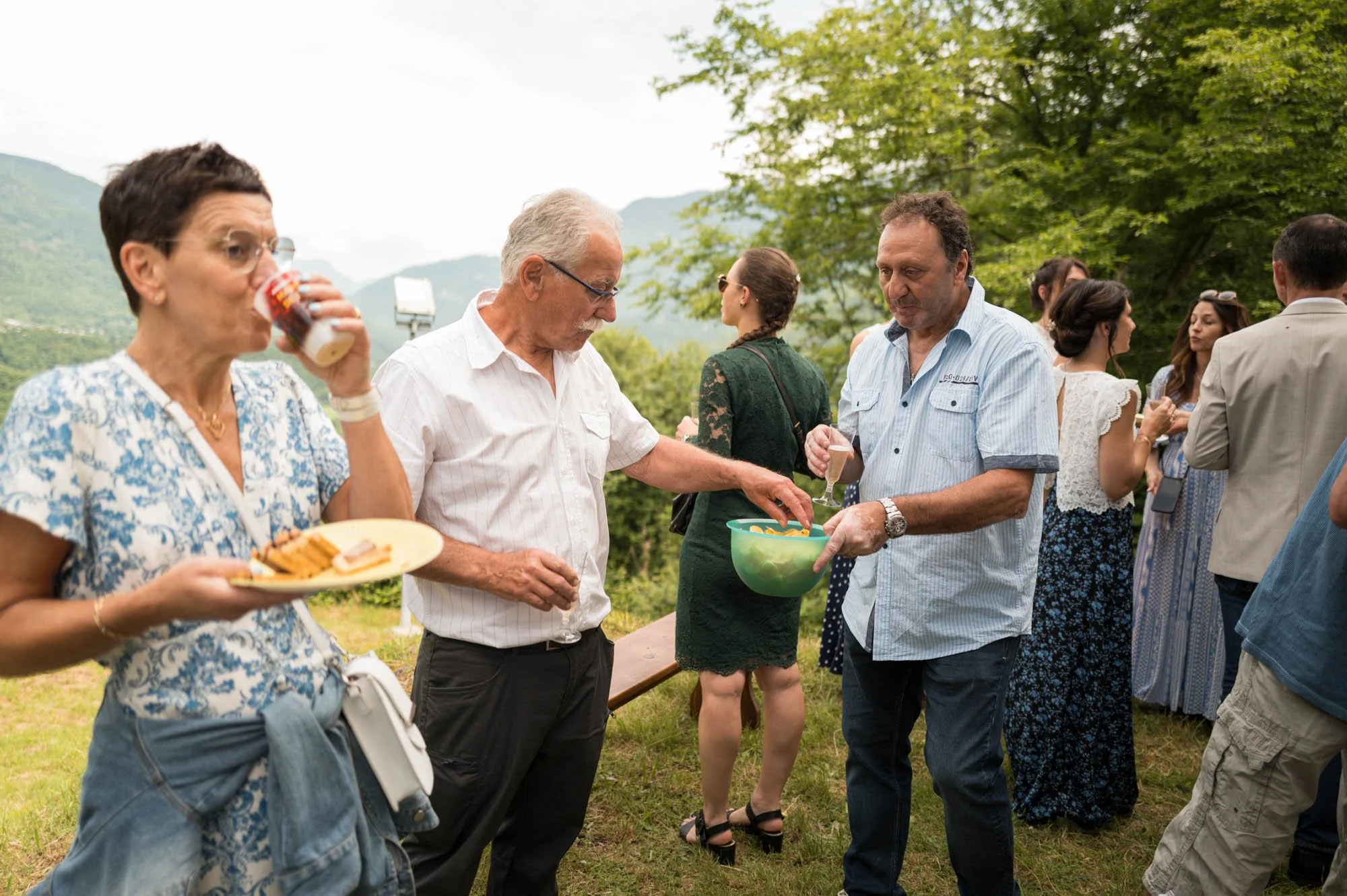 Groupe de personnes lors d'une fête en plein air, some mangeant et d'autres discutant, dans un cadre naturel avec des montagnes en arrière-plan.
