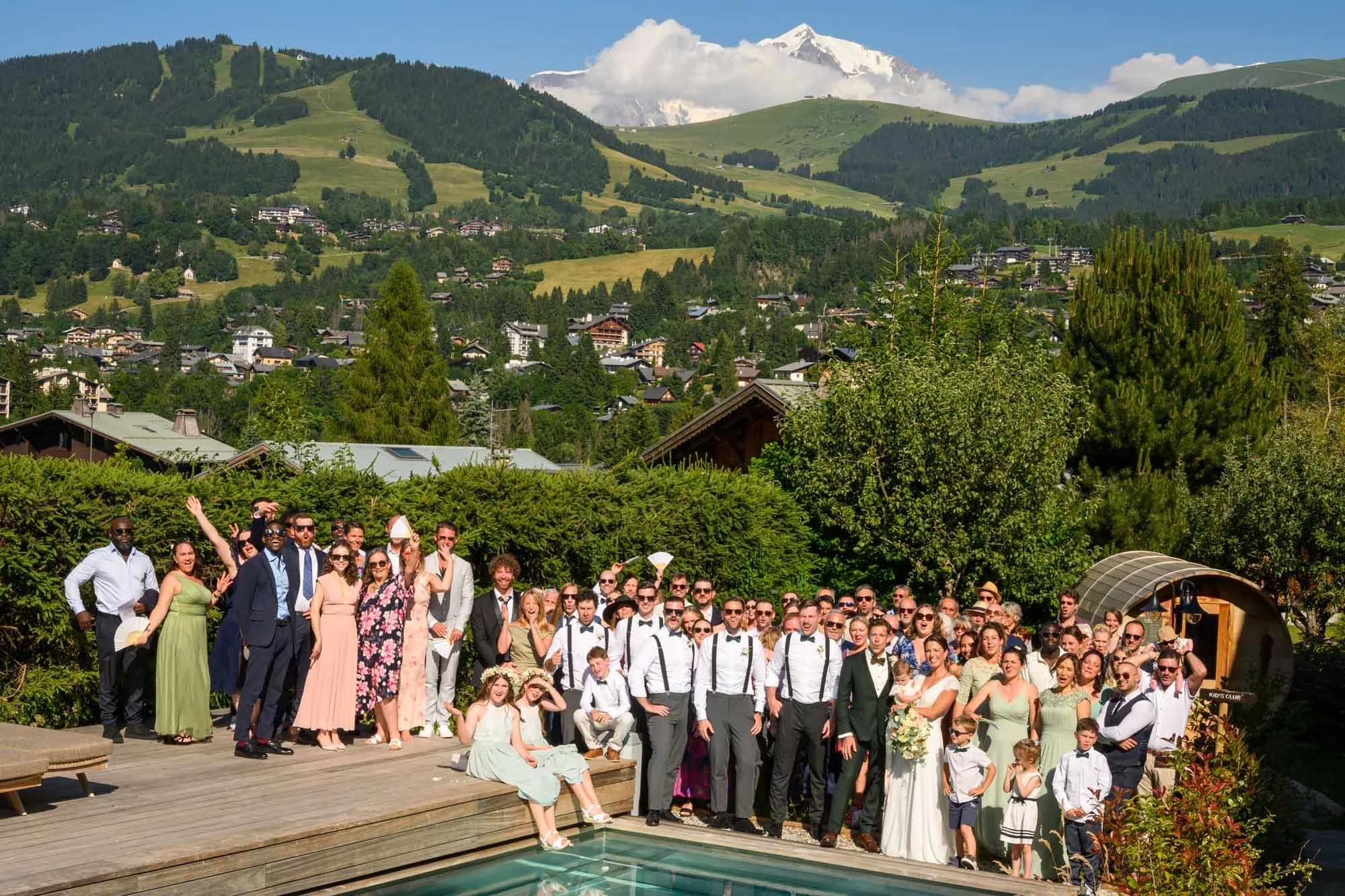 Groupe de personnes lors d'un mariage en plein air, avec montagnes en arrière-plan, soleil, arbres et piscine.