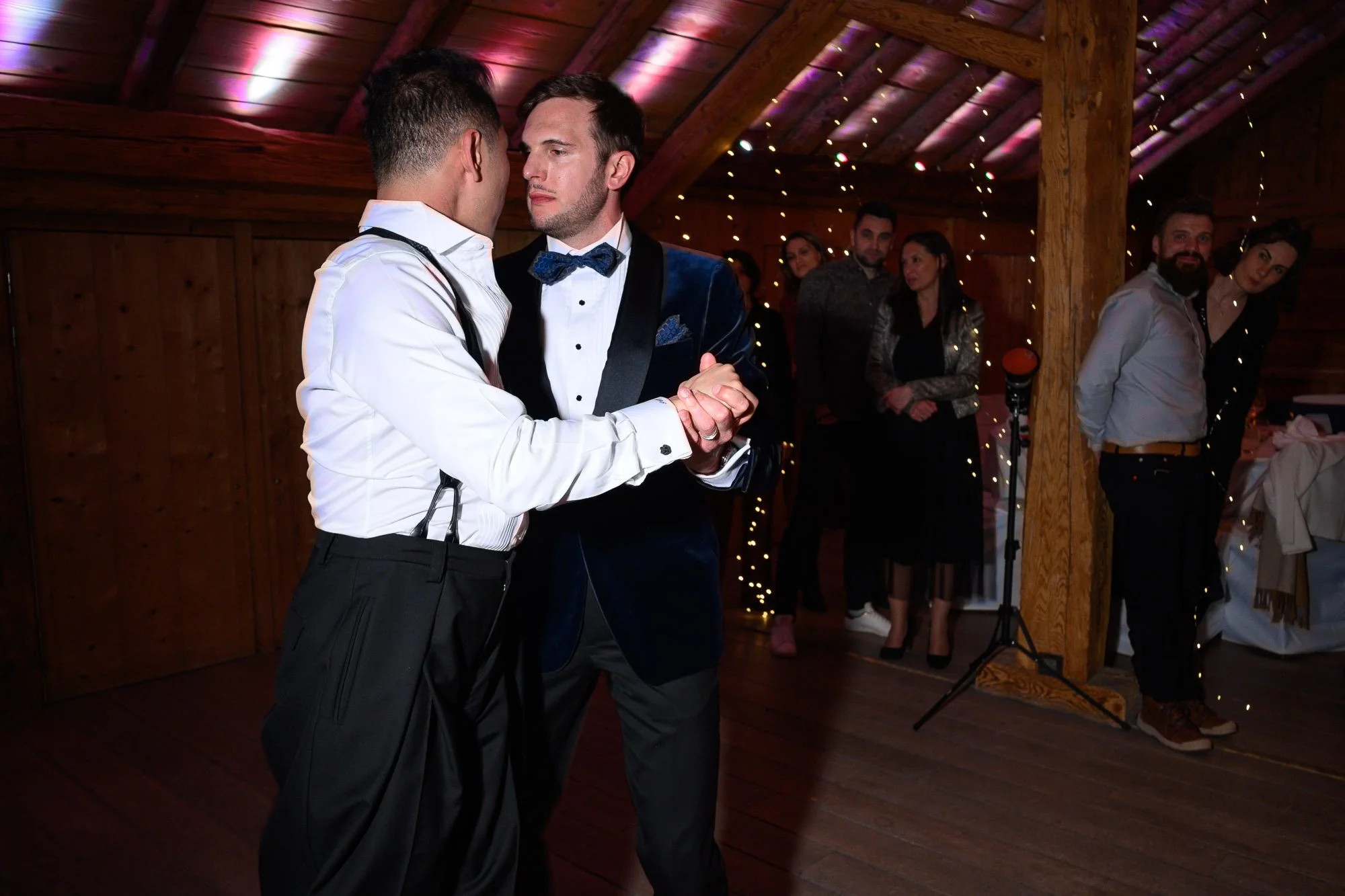 Deux hommes dans une soirée dansante, un d'eux porte une veste en velours bleu et un nœud papillon, l'autre une chemise blanche avec des bretelles, ils dansent ensemble. En arrière-plan, plusieurs personnes regardent et discutent dans une pièce en bo