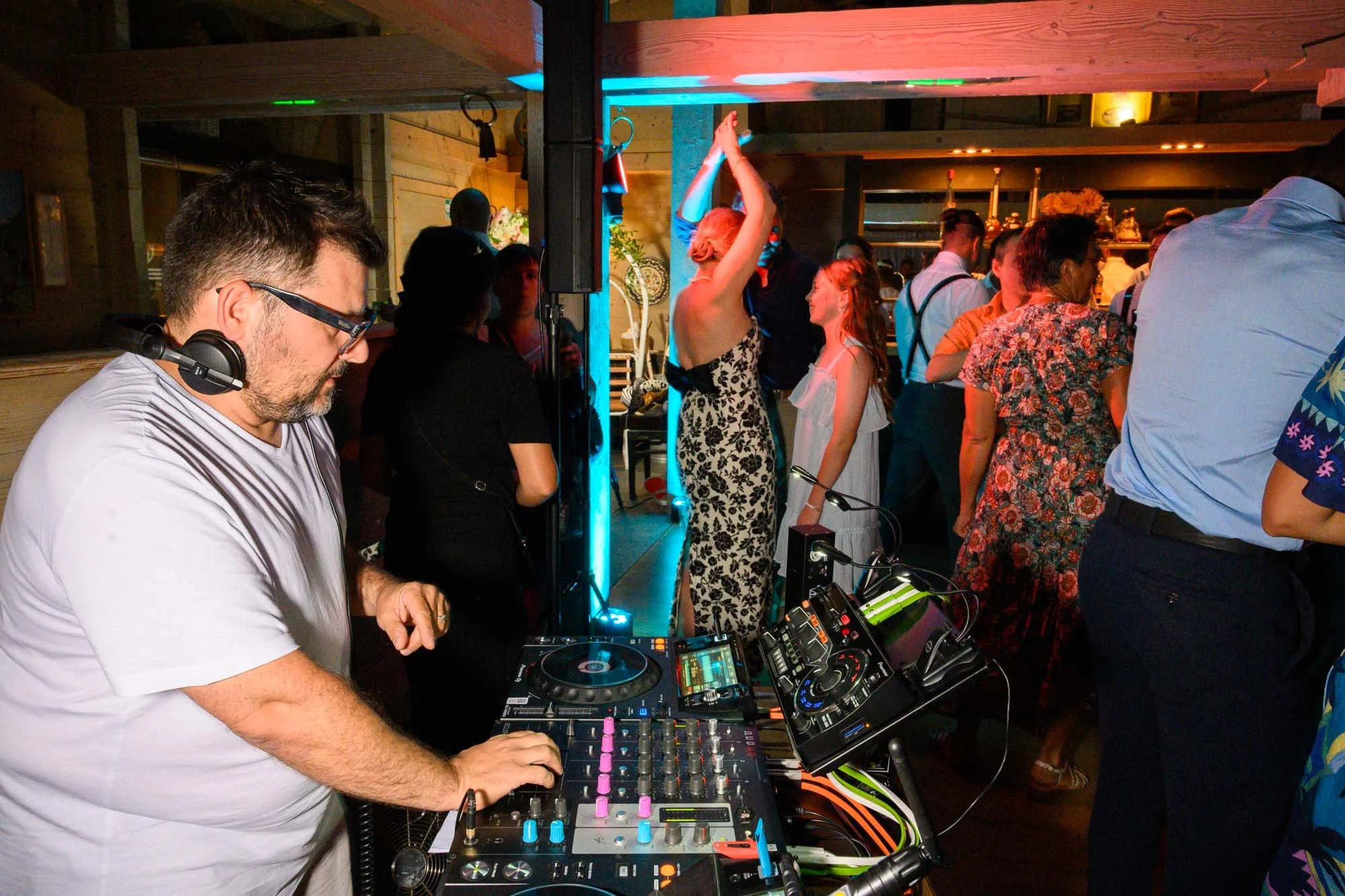 Un DJ avec un casque, utilisant du matériel de mixage, dans un lieu où des personnes dansent et socialisent, ambiance de fête