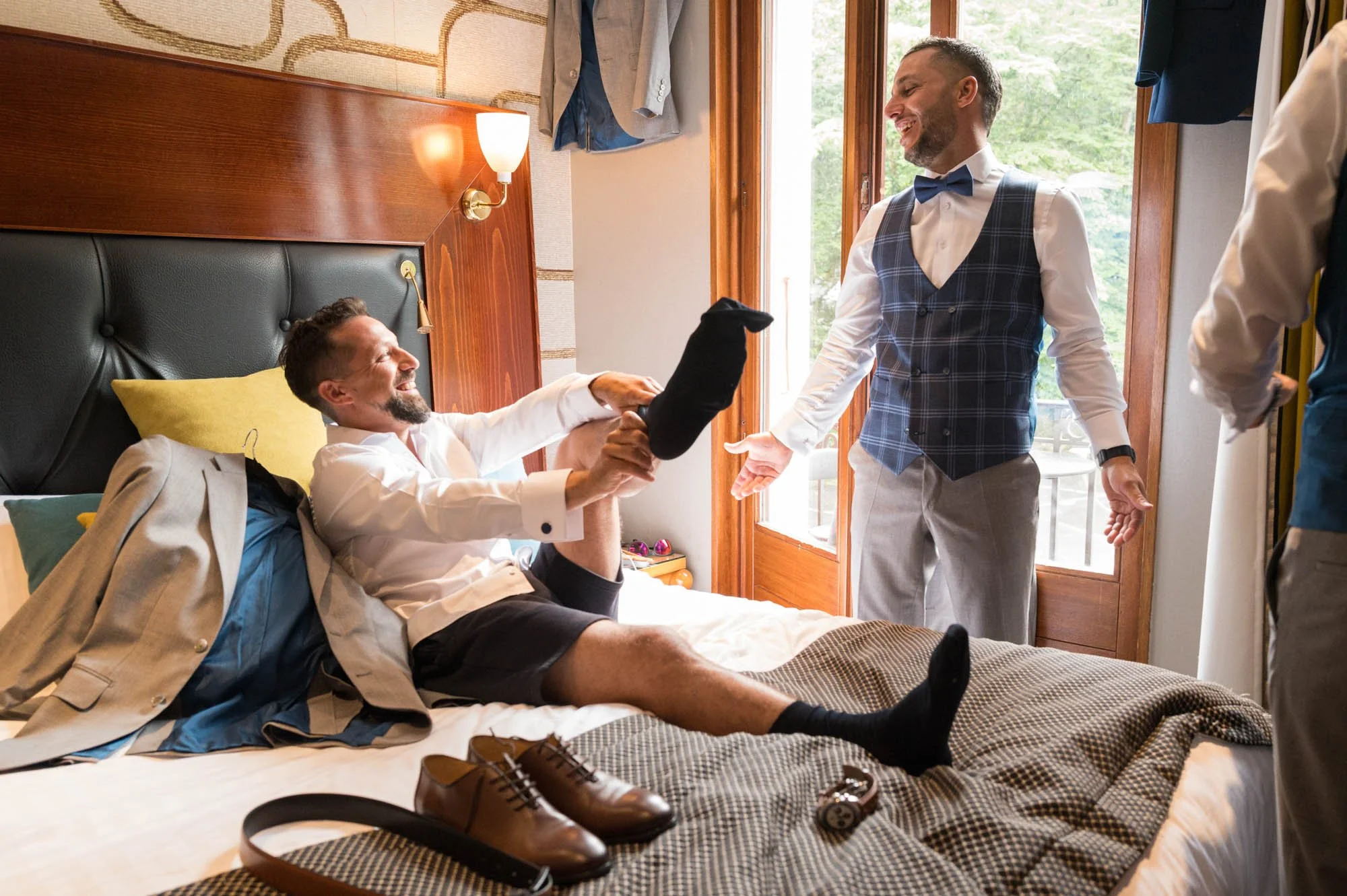 Deux hommes dans une chambre en train de jouer, l'un assis sur le lit avec un costume posé à côté, l'autre debout, tous deux souriants, avec des vêtements formels.