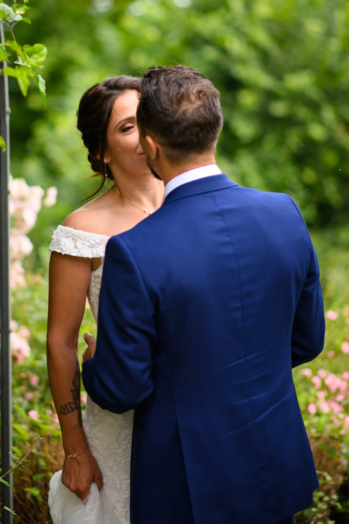 Un couple lors d'une séance photo de mariage dans un jardin, l'homme en costume bleu foncé et la femme en robe de mariage blanche, face à face, entourés de verdure.