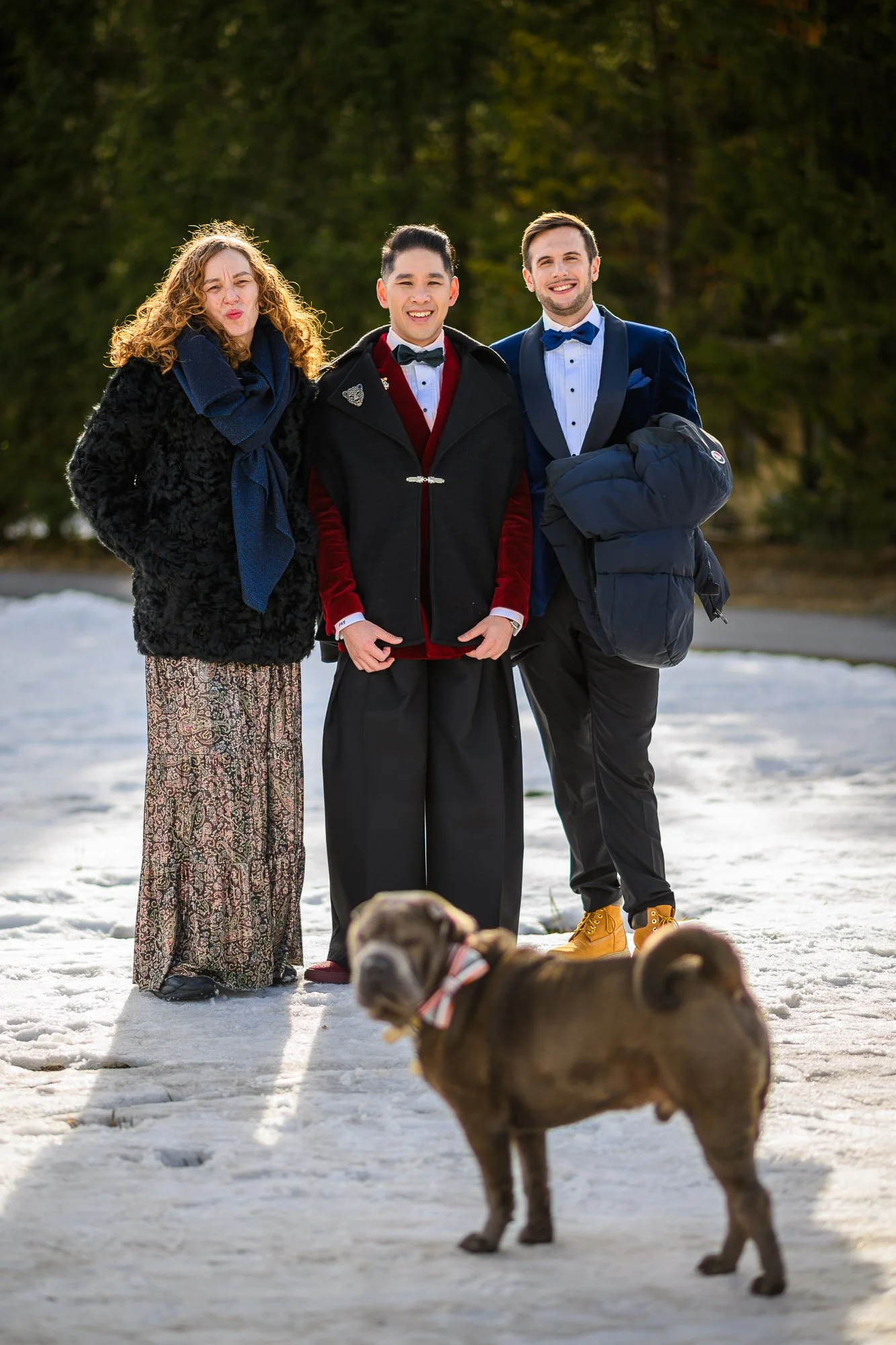 Trois personnes habillées élégamment pour une occasion spéciale, posant à l'extérieur sur la neige avec un chien en premier plan