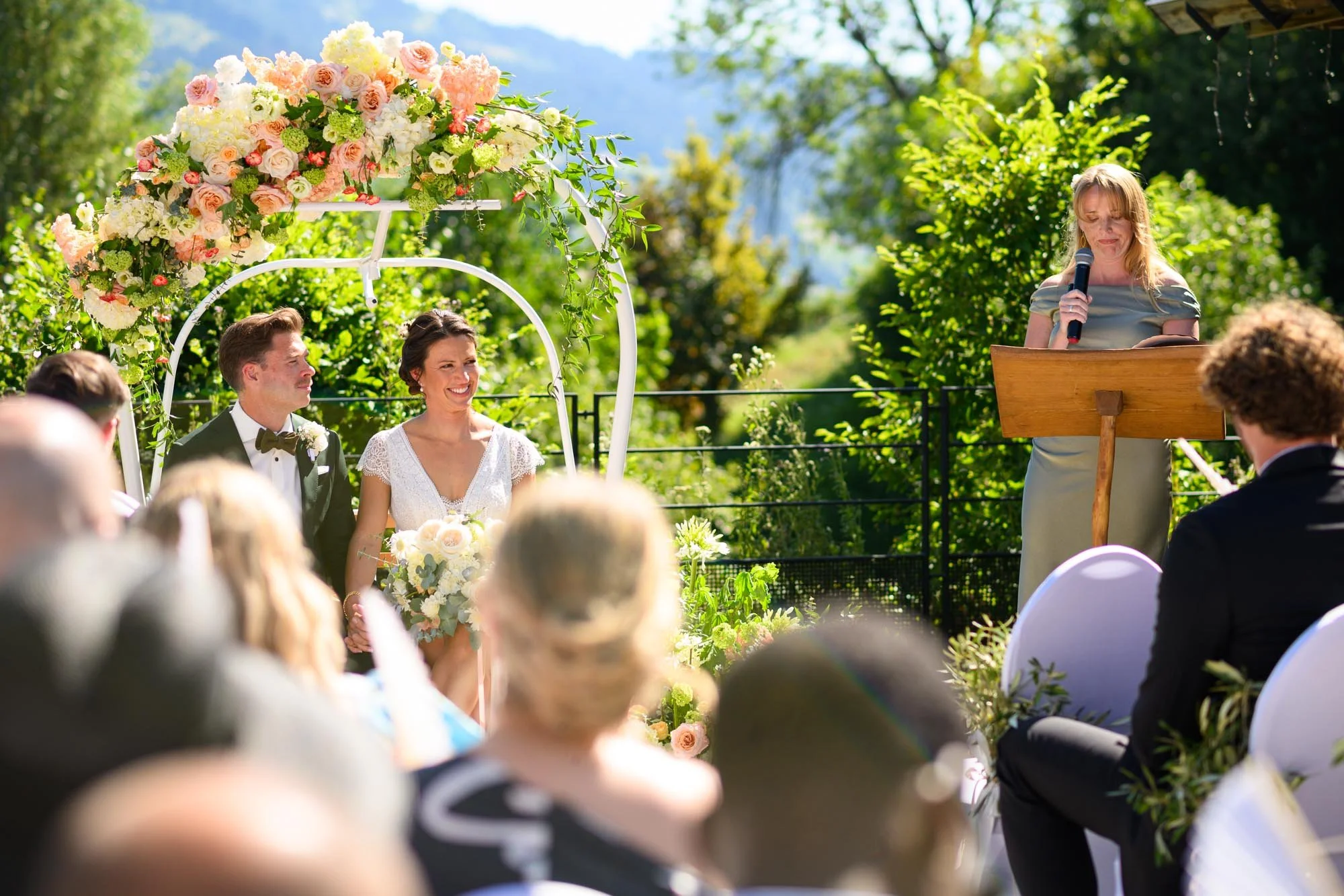 Un mariage en plein air avec un couple assis sous une arche de fleurs roses et blanches, et une femme en train de lire ou de prononcer un discours. Les invités sont assis en face, regardant vers la scène