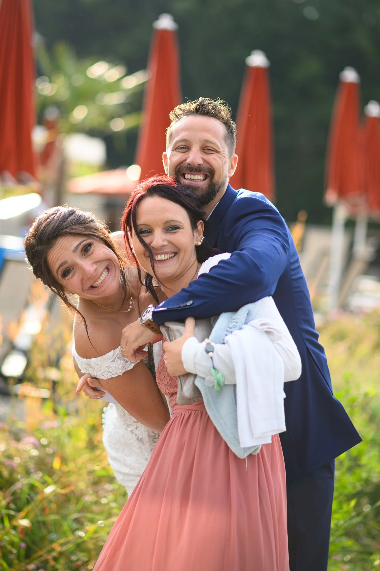 Trois personnes souriantes en plein câlin en extérieur, avec des parasols orange en arrière-plan et un jardin verdoyant.