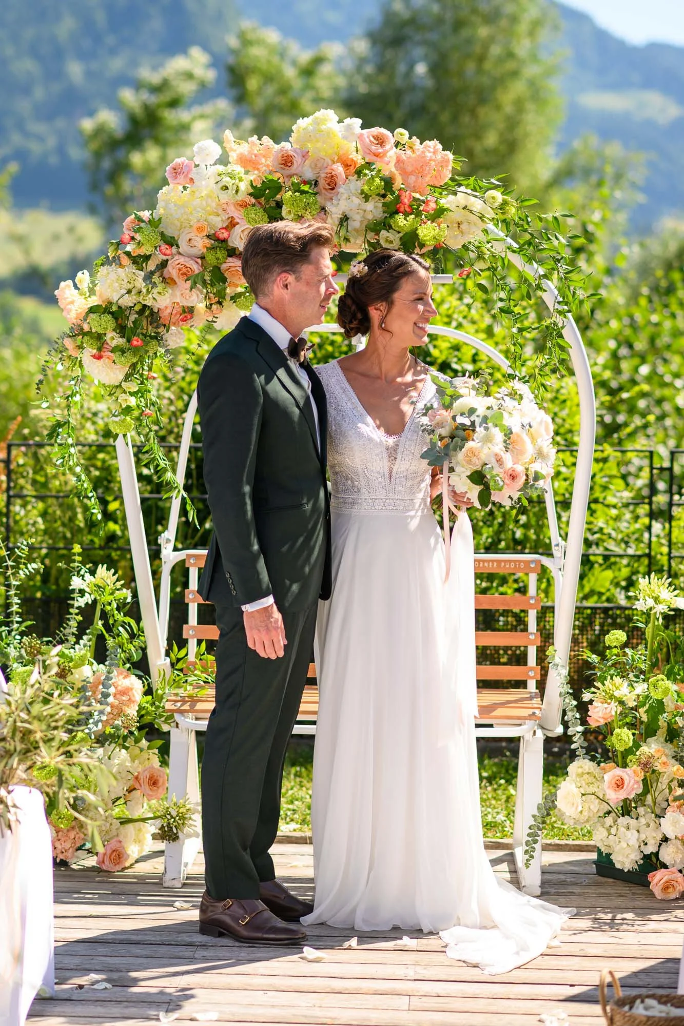 Un couple de mariés, homme en costume noir et femme en robe blanche, se tiennent près d'une arche florale lors d'une cérémonie de mariage extérieure en plein air, avec un décor de fleurs et une nature verdoyante en arrière-plan.