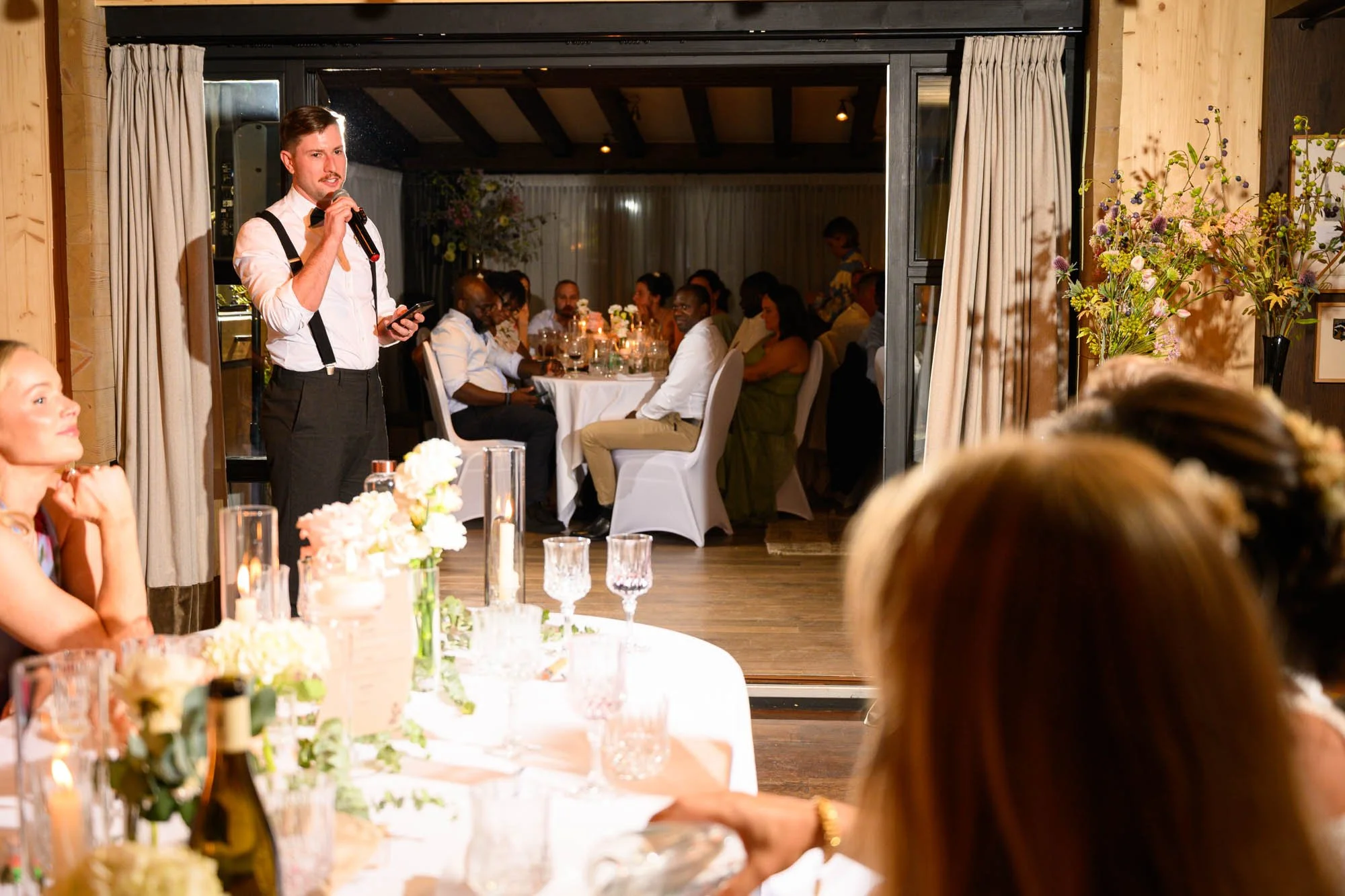 Un homme parlant au micro lors d'un événement dans une salle de réception, avec des invités assis à une table décorée avec des fleurs et des bougies.