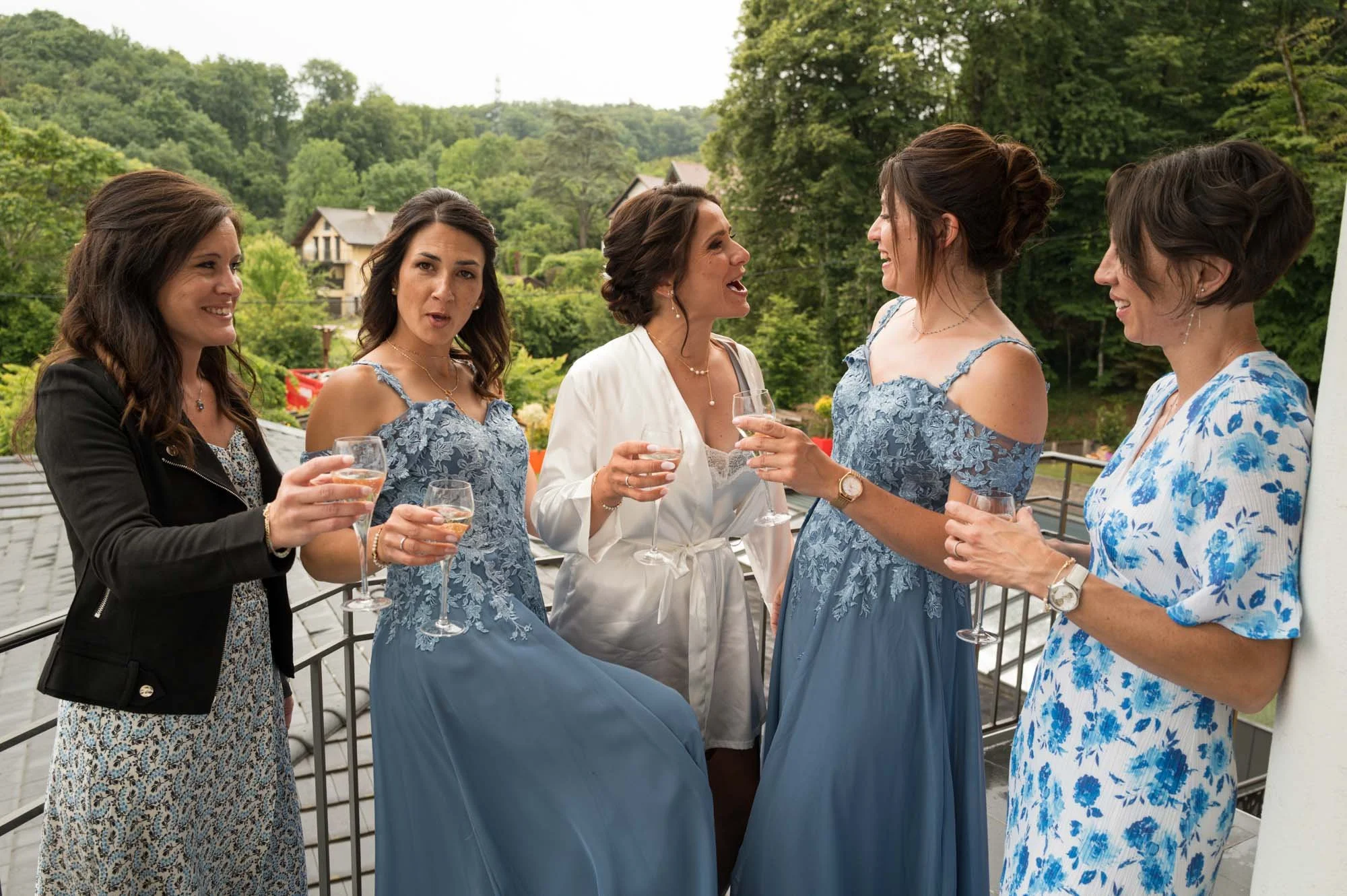Groupe de six femmes discutant et levant des verres lors d'une fête en extérieur sur un balcon avec vue sur une forêt verte.
