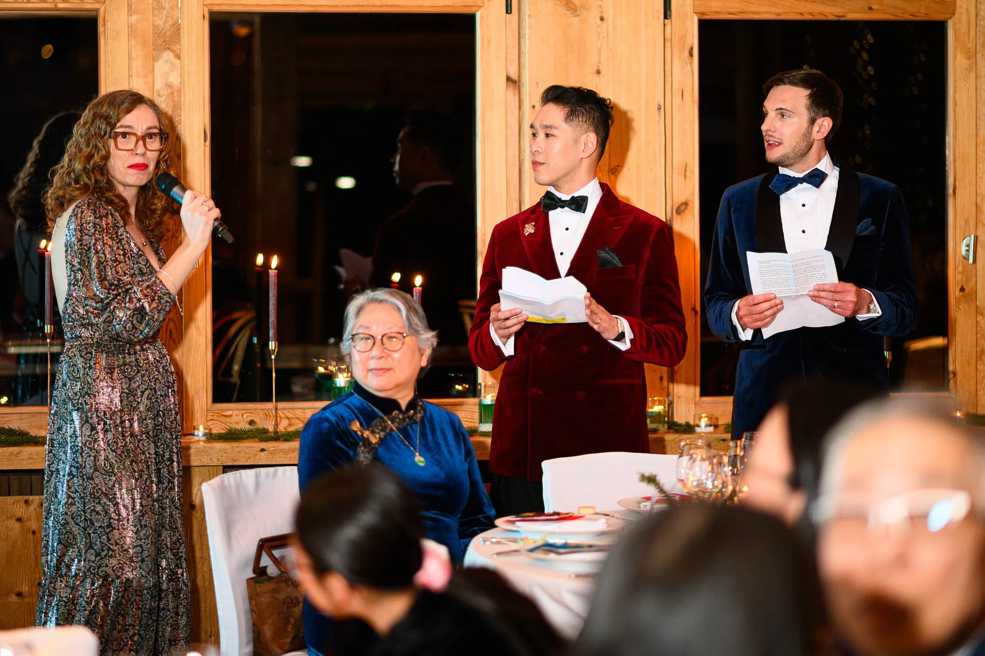Un groupe de personnes lors d'une cérémonie ou d'un repas, avec une femme parlant au microphone à gauche, deux hommes en costumes parlant à droite, et une femme assise en bleu au centre, dans une salle avec décorations chaleureuses.