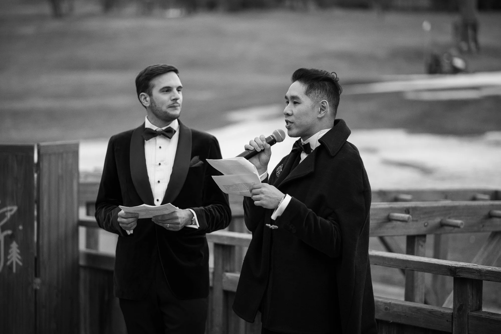 Deux hommes en costume, l'un parlant dans un microphone, lors d'une cérémonie en plein air.