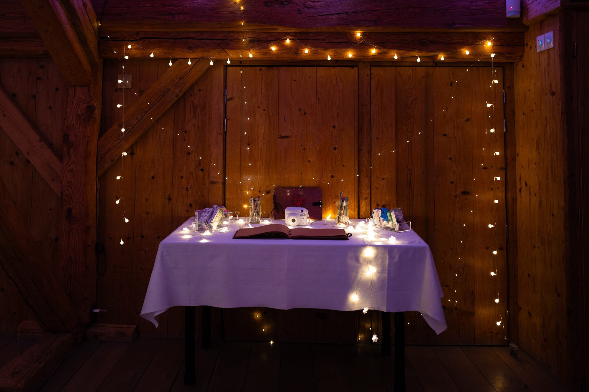 Décor de fête avec une table recouverte d'une nappe blanche, guirlandes lumineuses et quelques objets décoratifs, dans une pièce en bois.