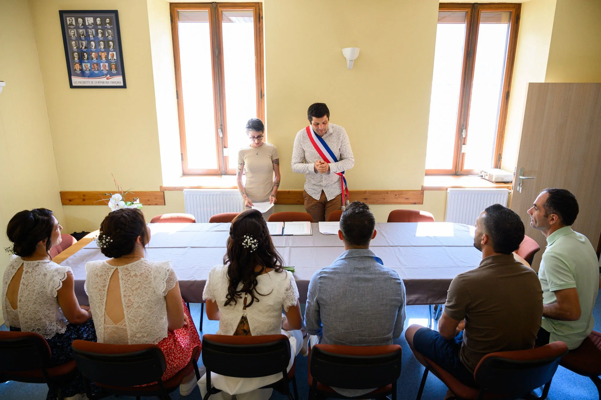 Un groupe de personnes assises autour d'une table, lors d'une cérémonie de mariage, avec deux personnes debout, dont un homme portant une sash tricolore, recueillant des vœux ou prononçant un discours, dans une pièce lumineuse avec fenêtres et un tab
