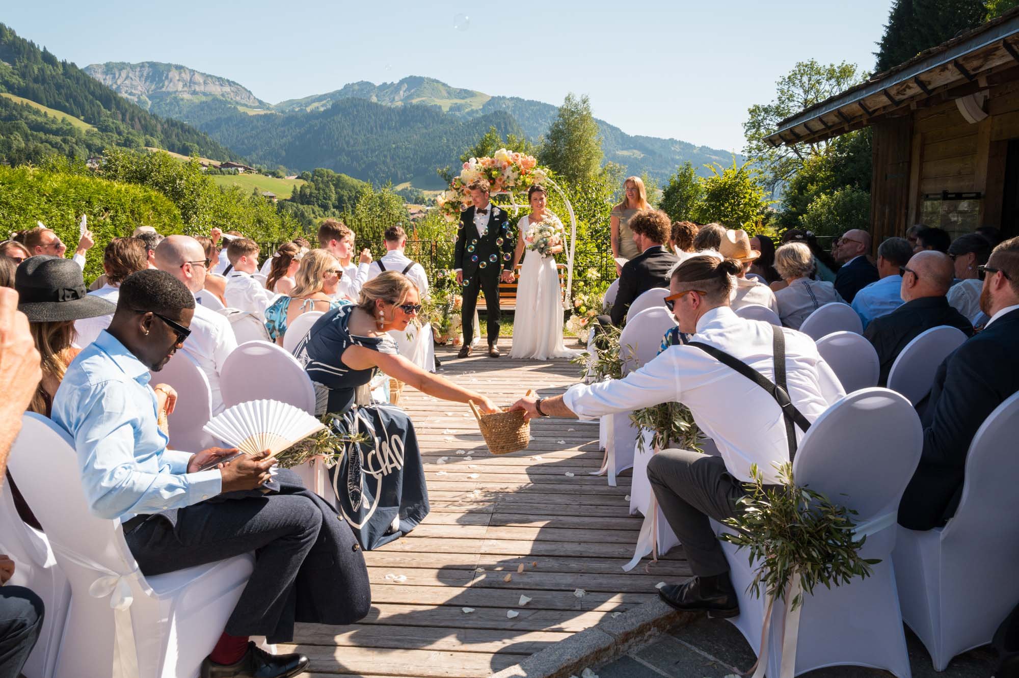 Un mariage en plein air avec un couple de mariés marchant sur une allée en bois entourée d'invités assis, dans un paysage montagnard ensoleillé.