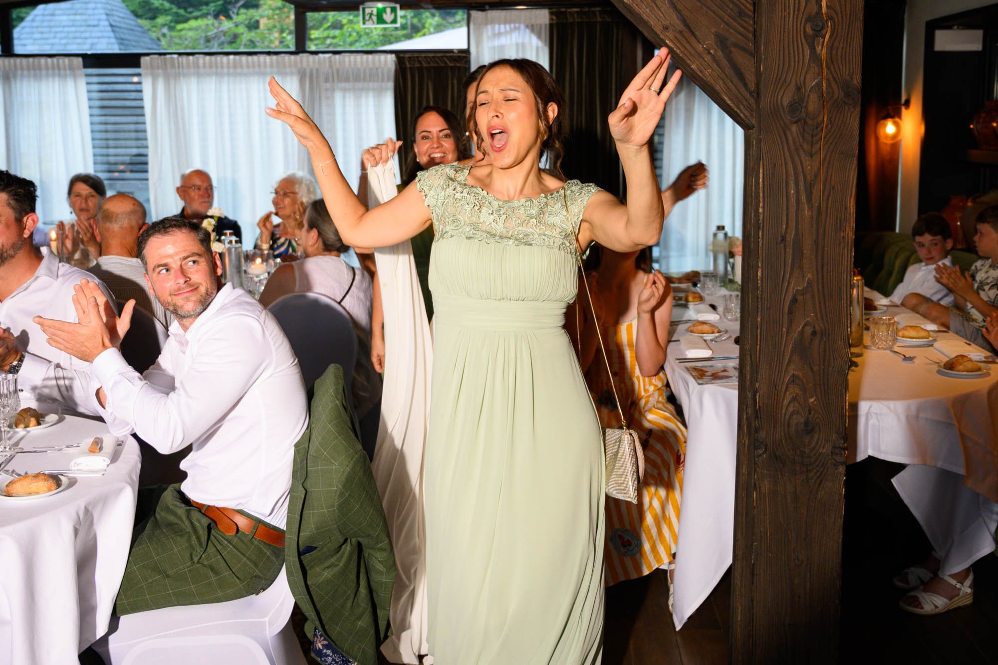 Une femme en robe verte chante ou danse avec les yeux fermés lors d'un rassemblement social, entourée de personnes assises à une table de restaurant ou de salle de fête.