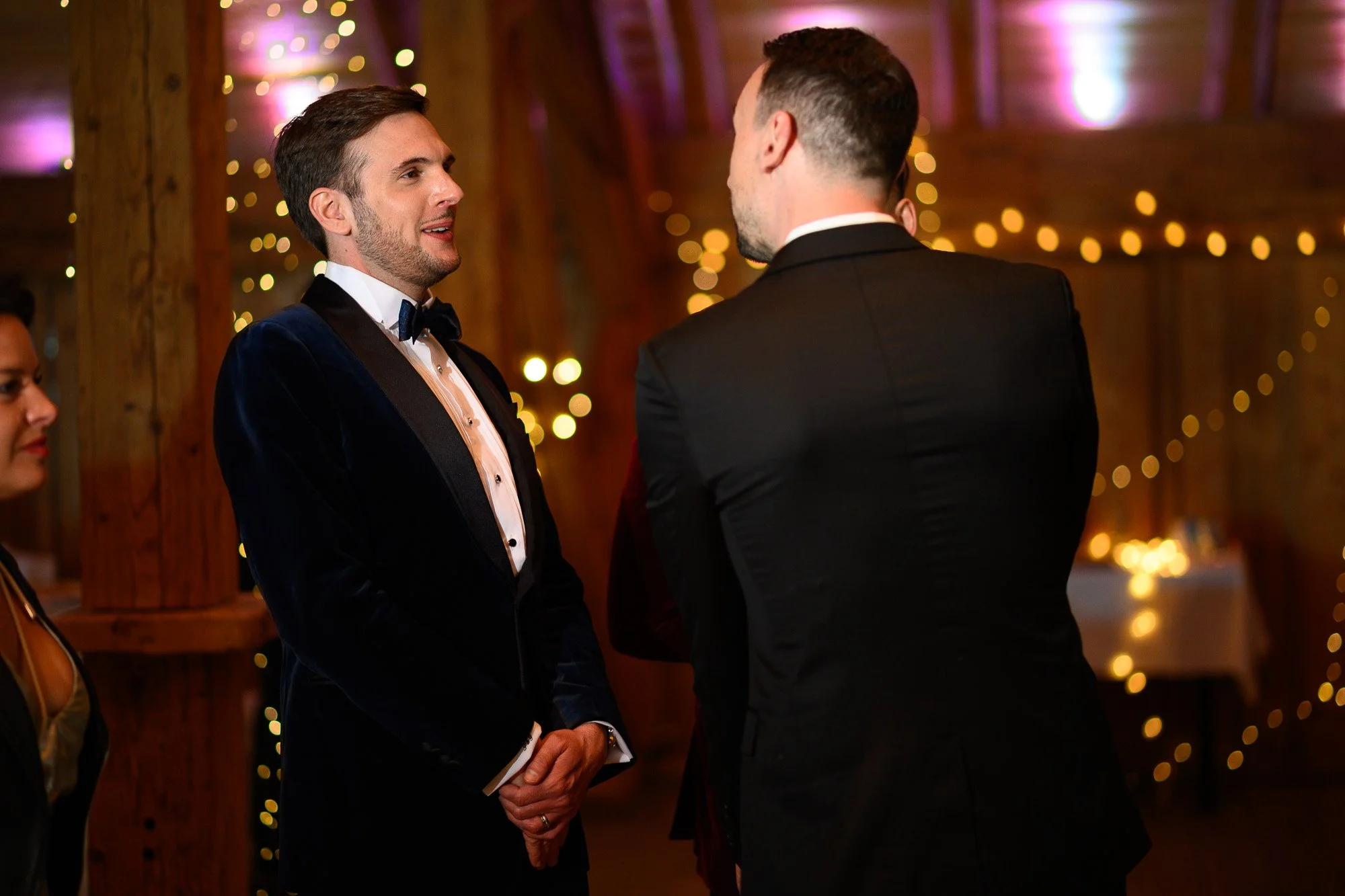 Deux hommes en veste et cravate discutent lors d'une célébration dans une salle décorée avec des lumières chaleureuses et des guirlandes lumineuses en arrière-plan.