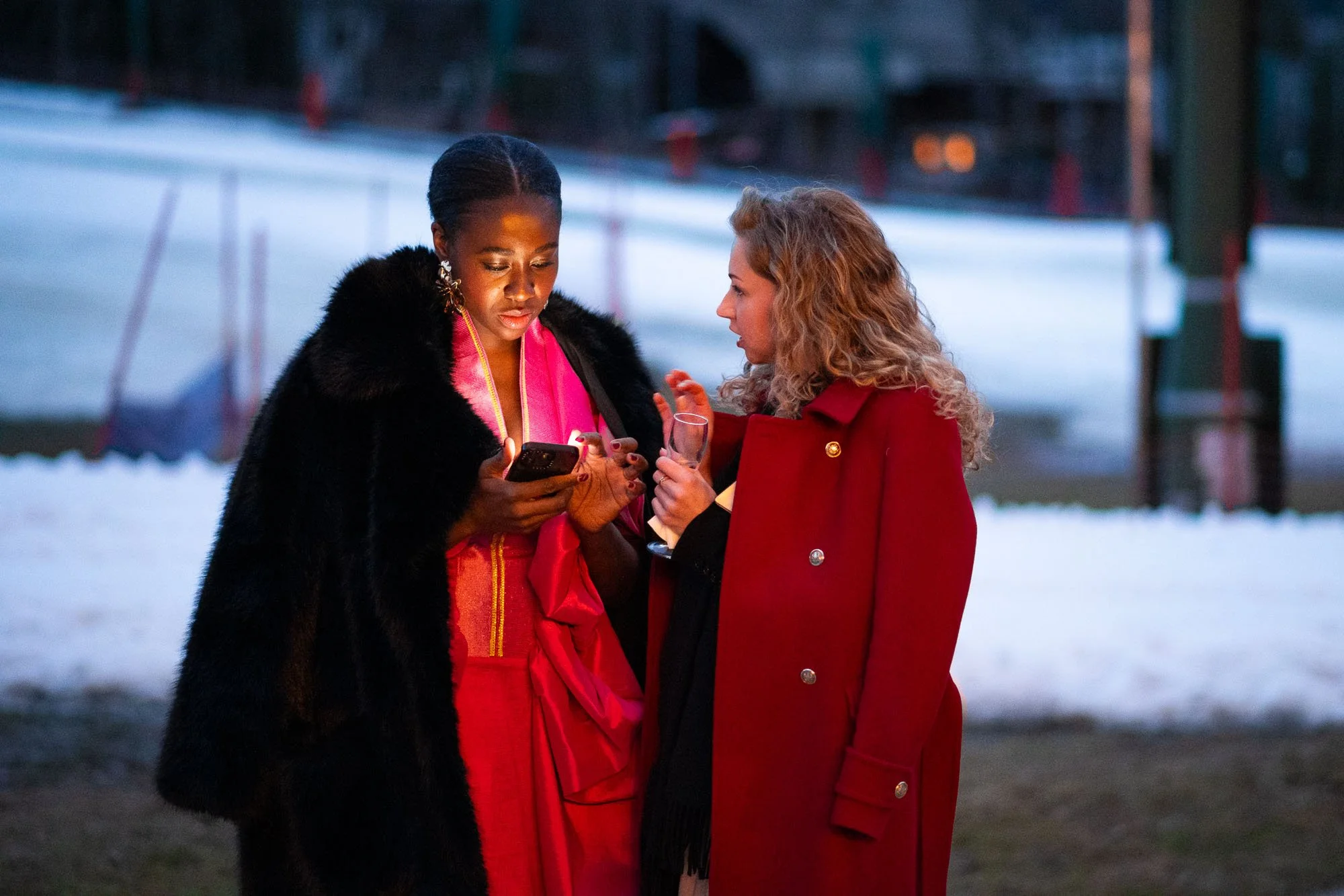 Deux femmes vêtues d'extravagantes tenues colorées regardent un téléphone portable, semblant discuter dans un lieu enneigé lors de la soirée.