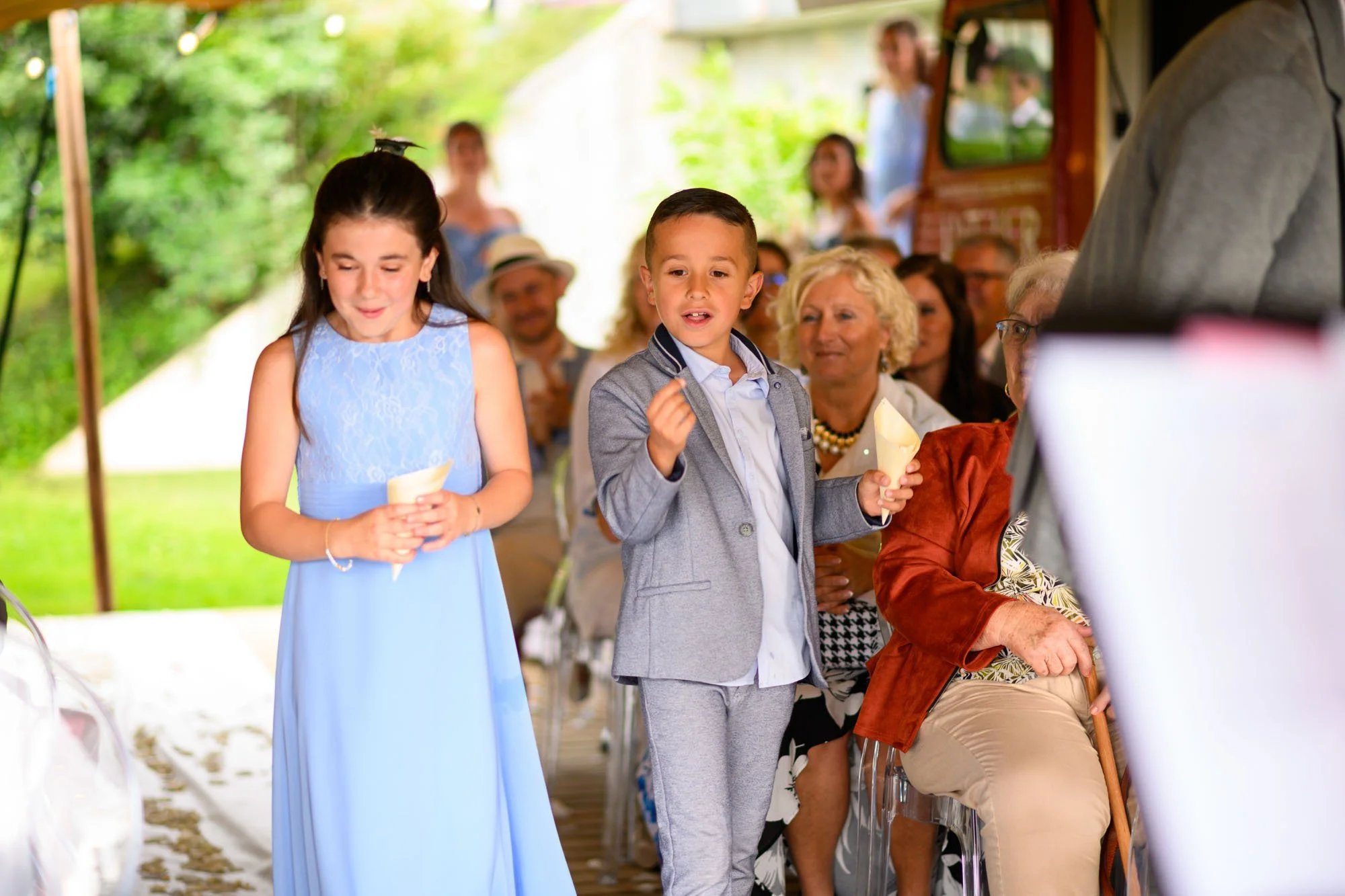 Deux enfants, une fille en robe bleue et un garçon en costume gris, tiennent des cônes de glace lors d'une fête en plein air avec des invités assis autour.
