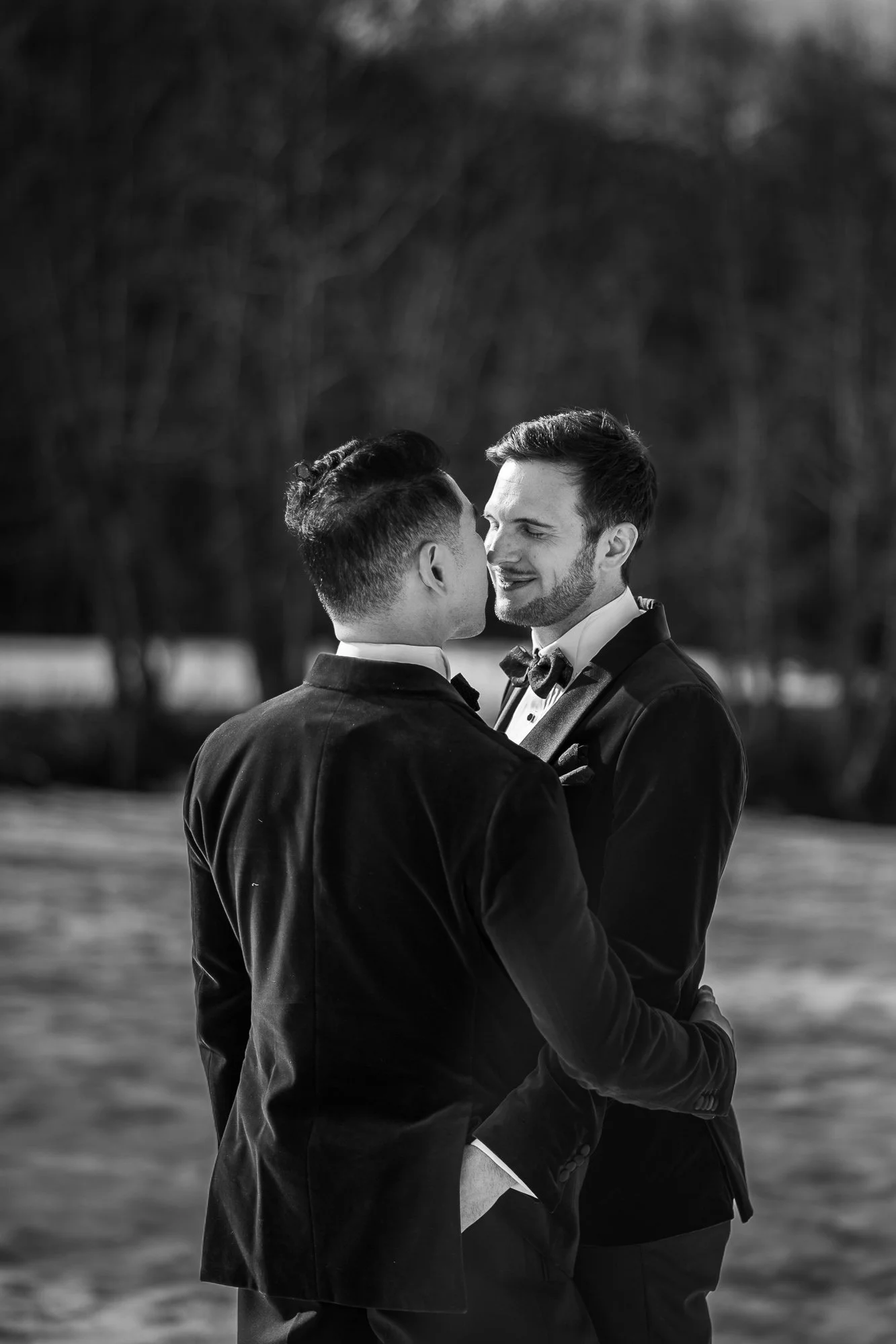 Deux hommes en costume se tiennent proches, dans un cadre extérieur, en train de s'embrasser tendrement, probablement lors d'un mariage ou d'une occasion spéciale.