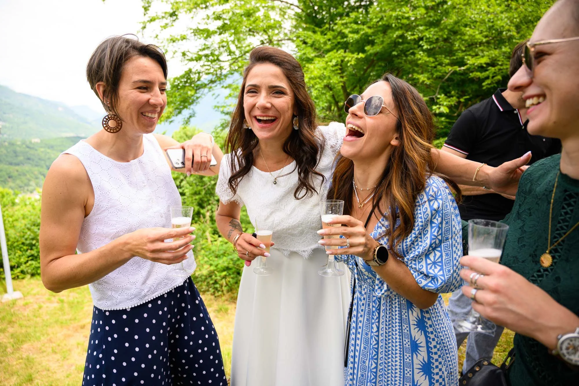 Groupe de femmes souriantes lors d'une célébration en plein air, tenant des verres de boisson, avec des arbres verts en arrière-plan.