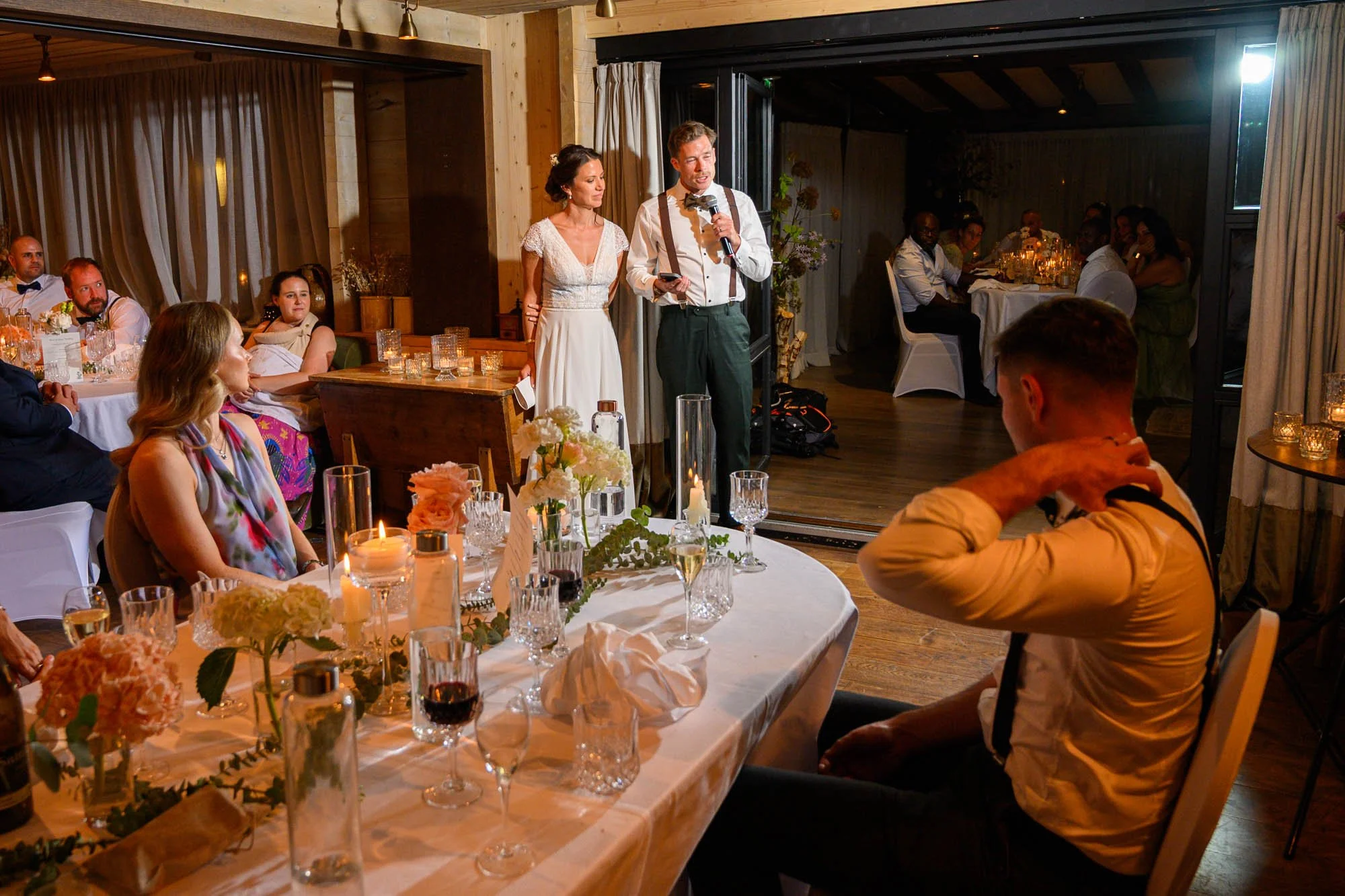 Un mariage dans une salle décorée avec des fleurs et des bougies, avec un couple marié parlant lors d'un discours, entouré d'invités assis à des tables. La scène est chaleureuse et élégante.