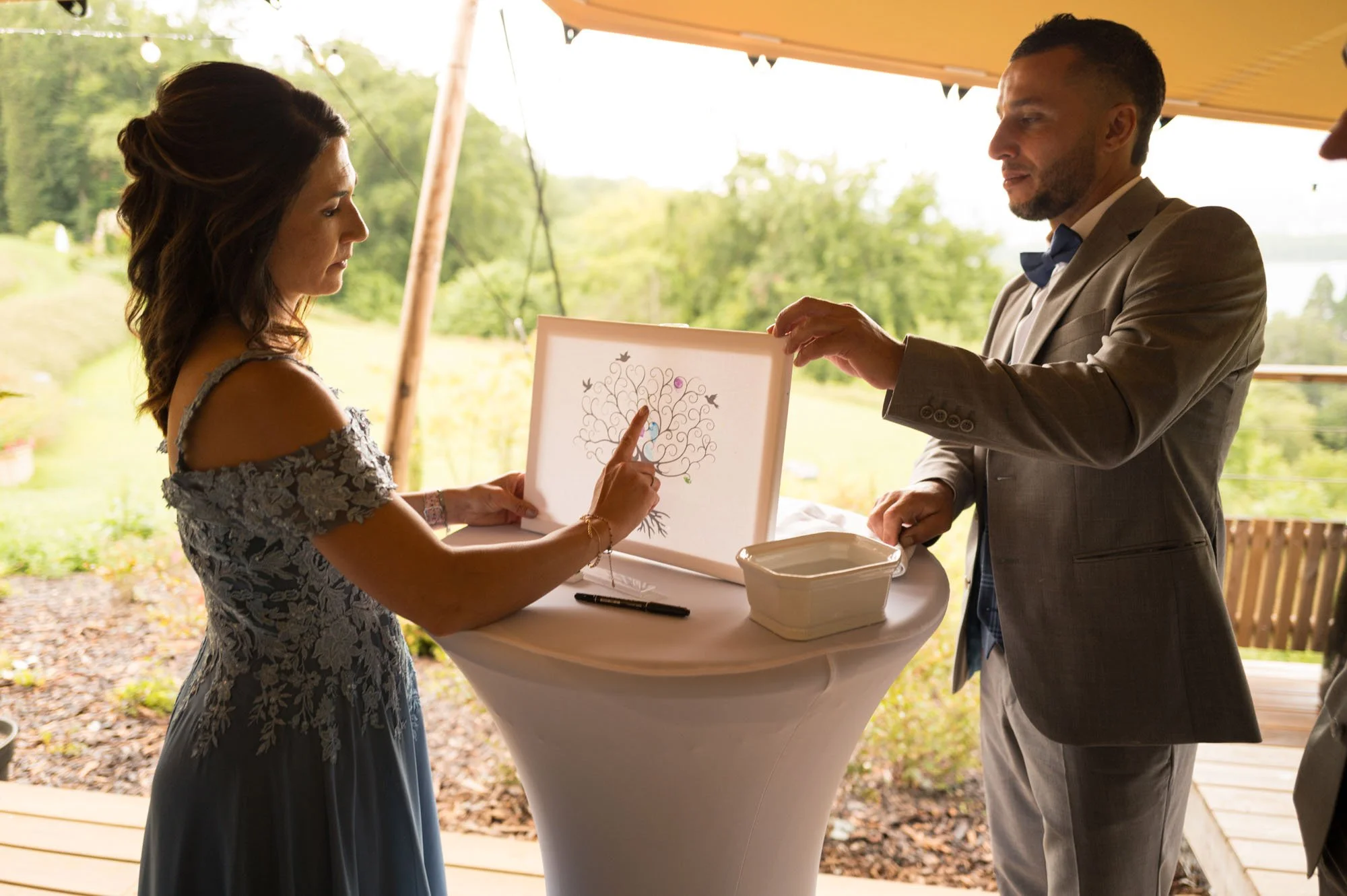 Un homme en costume gris et une femme en robe grise discutent autour d'une table haute à l'extérieur, avec un tableau illustrant un arbre au centre. L'environnement est verdoyant et ensoleillé.