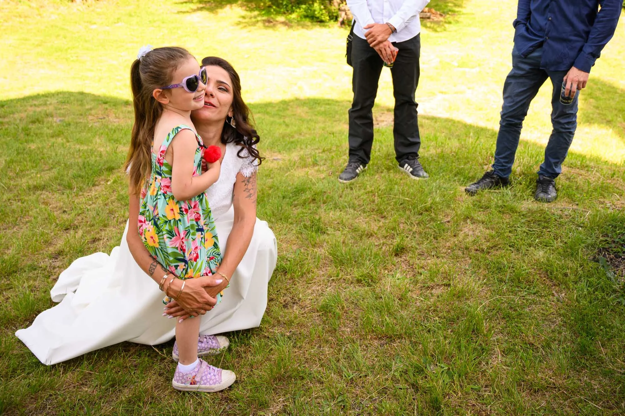 Une femme assise dans l'herbe, tenant une jeune fille portant des lunettes de soleil et une robe à fleurs, lors d'un événement en plein air, avec deux hommes en arrière-plan qui tiennent des boissons.
