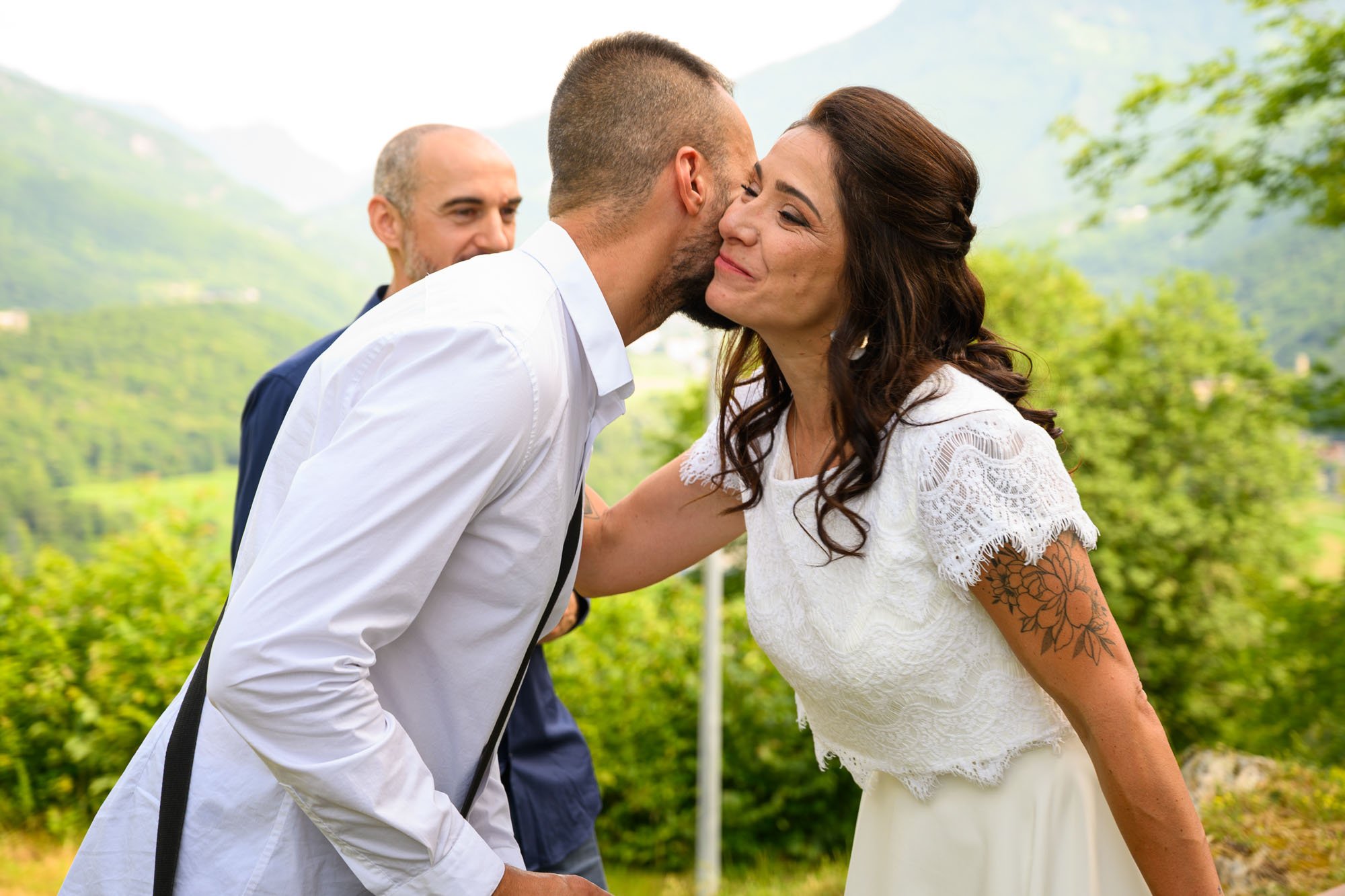 Un couple échange un baiser, un homme en chemise blanche et une femme avec une tatouage sur le bras, lors d'une cérémonie en plein air, avec un homme en costume en arrière-plan, dans un environnement naturel verdoyant.