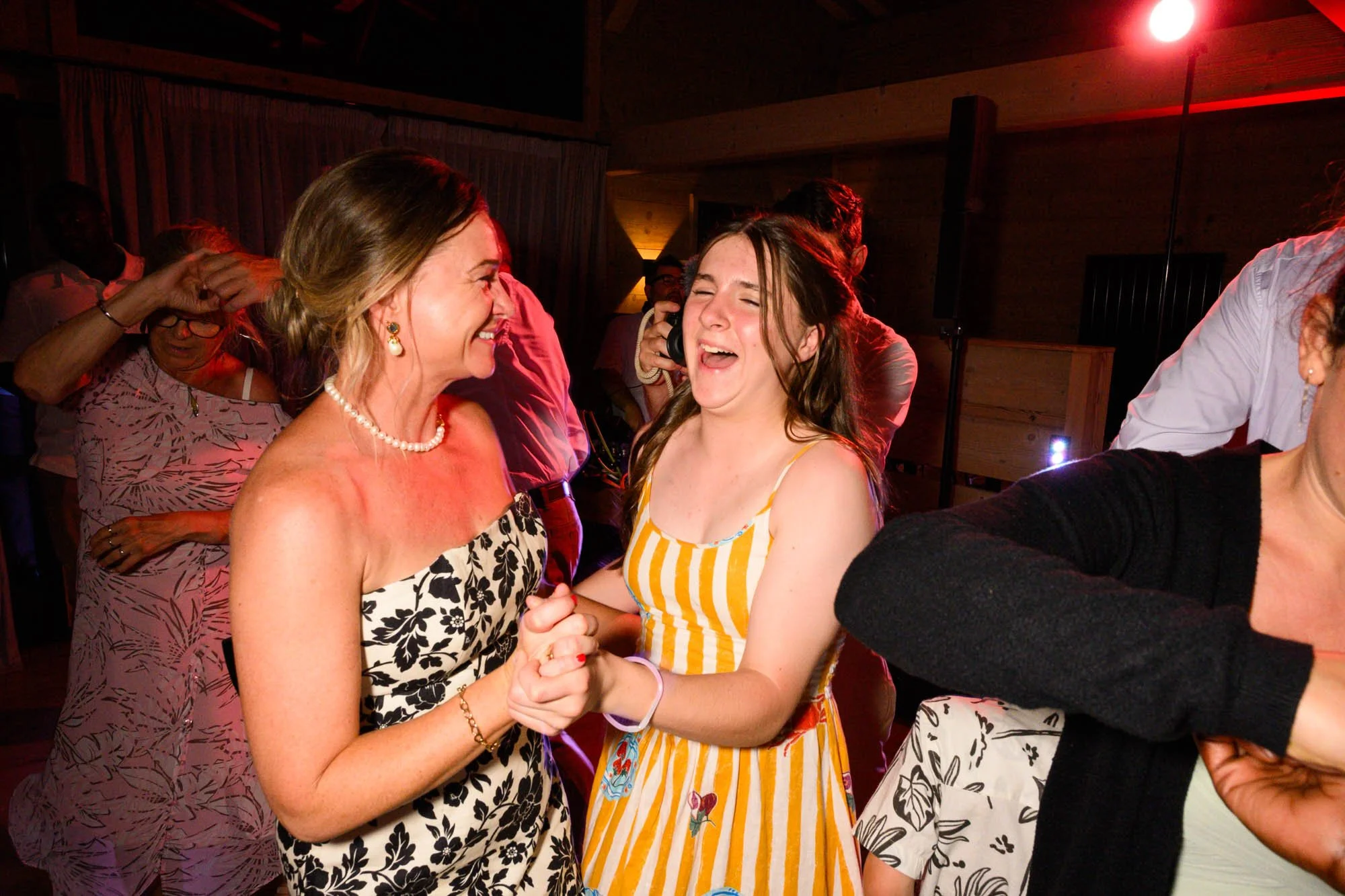 Deux femmes souriantes dansent et se tiennent la main lors d'une célébration dans une pièce éclairée par des lumières rouges et tamisées.