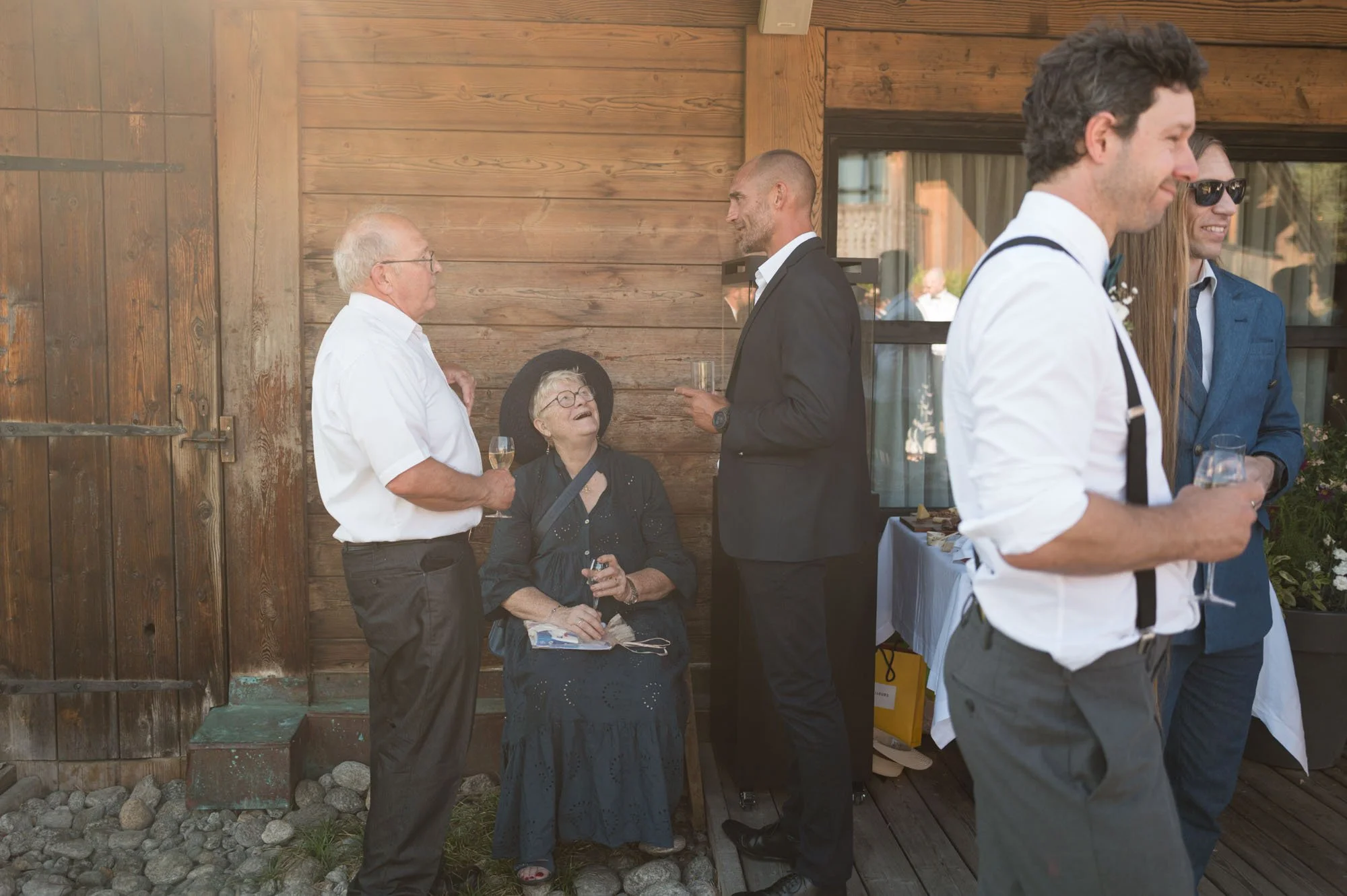 Groupe de personnes en train de discuter lors d'une réception, avec un fond en bois, plusieurs portes fenêtres et table de buffet à l'arrière.
