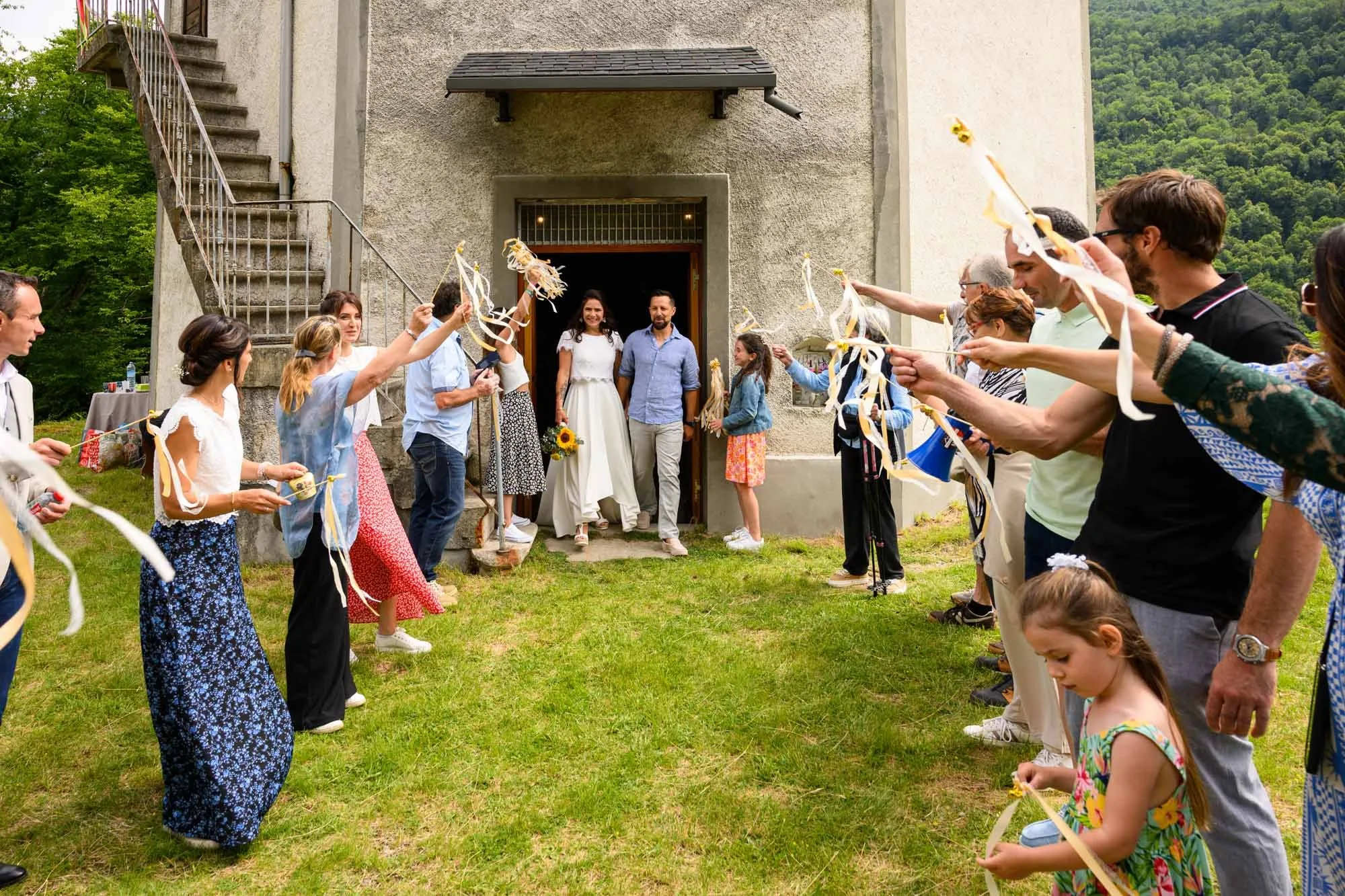 Groupe de personnes formant un couloir pour un mariage en extérieur, avec la mariée et le marié sortant de la bâtisse