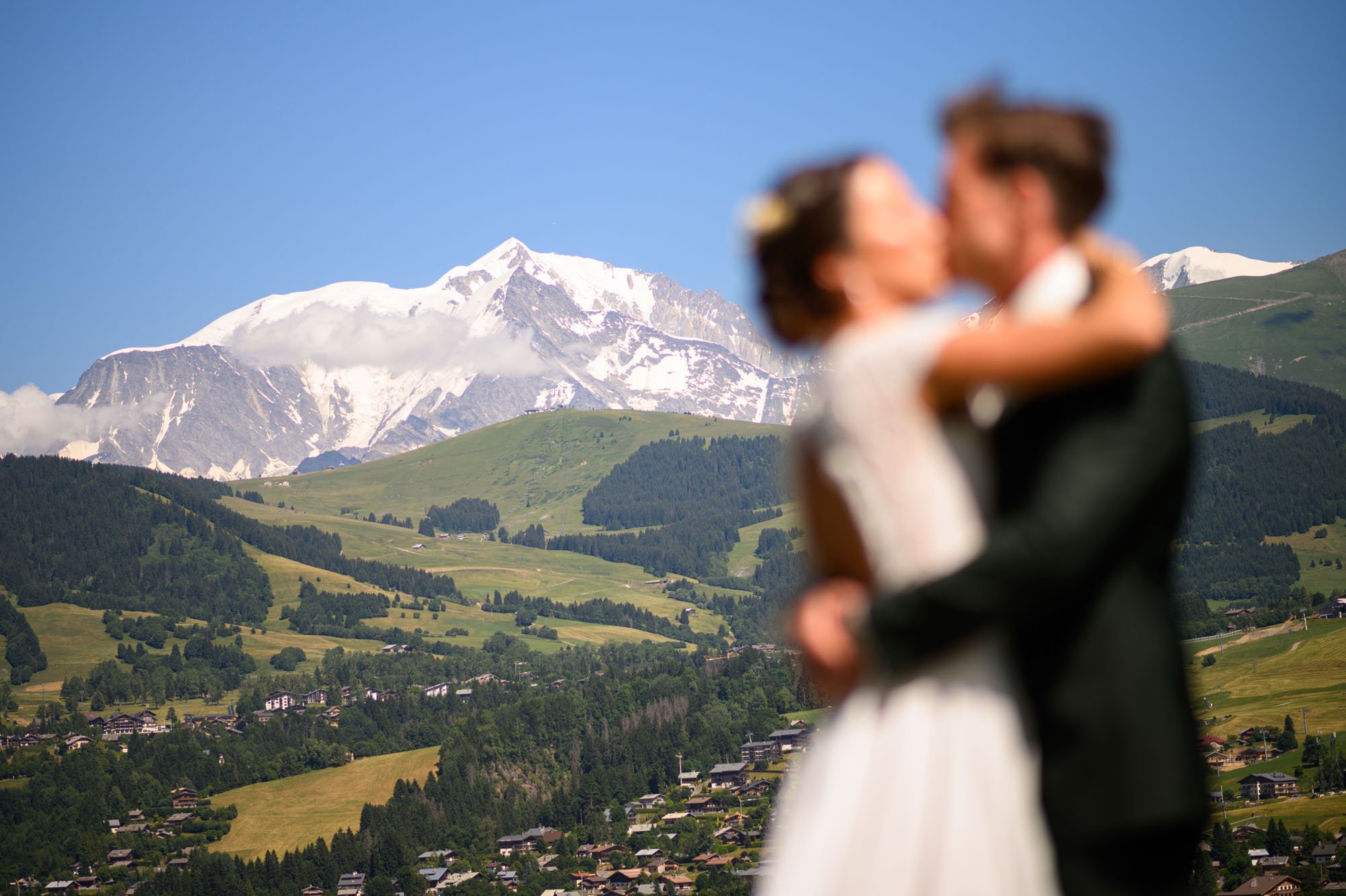 Un couple de mariés en gros plan dans une étreinte, avec un paysage de montagnes et de villages en arrière-plan.