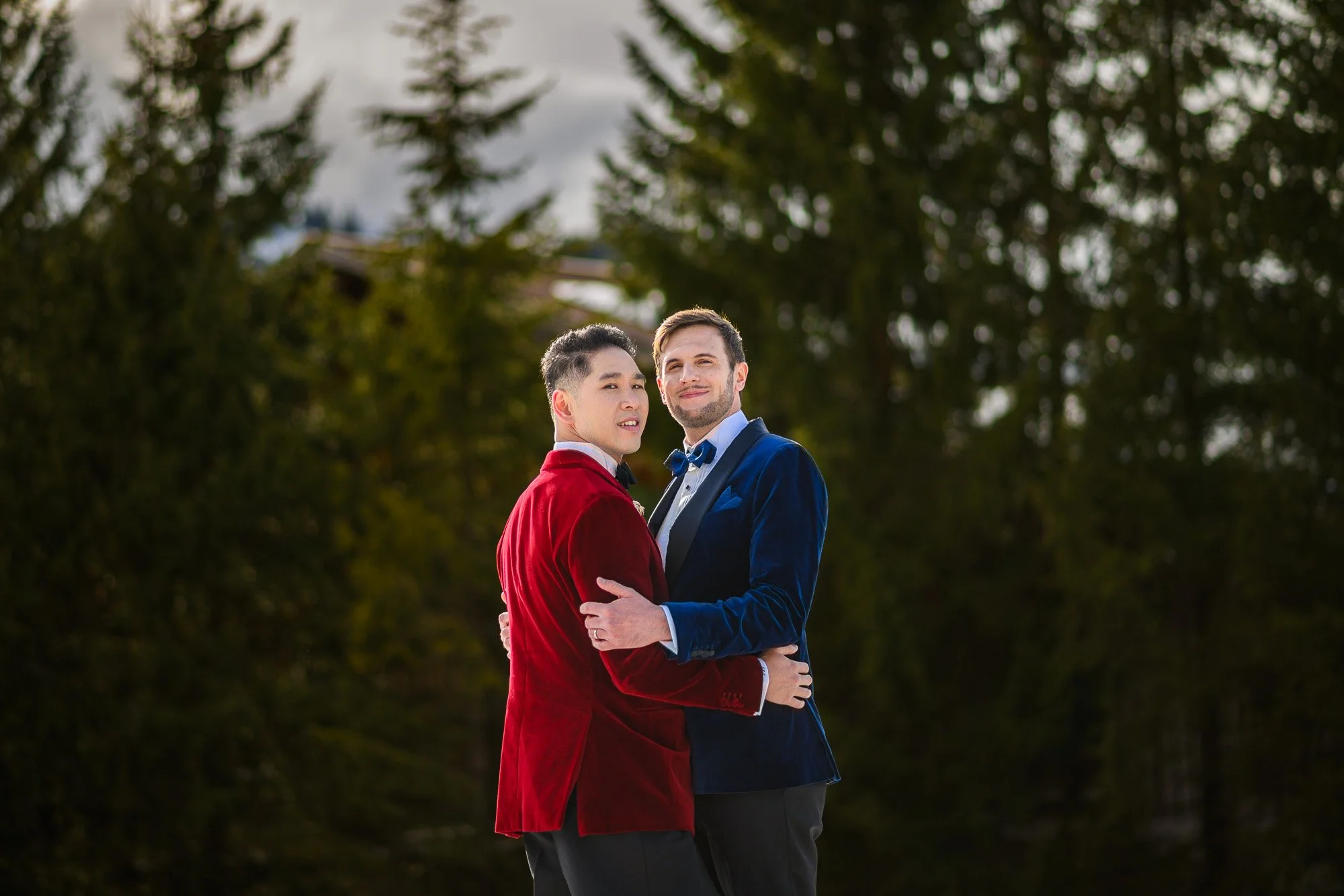 Deux hommes en costumes élégants, un en velours rouge et l'autre en velours bleu, en train de se tenir dans une nature verdoyante, partageant un moment d'affection.