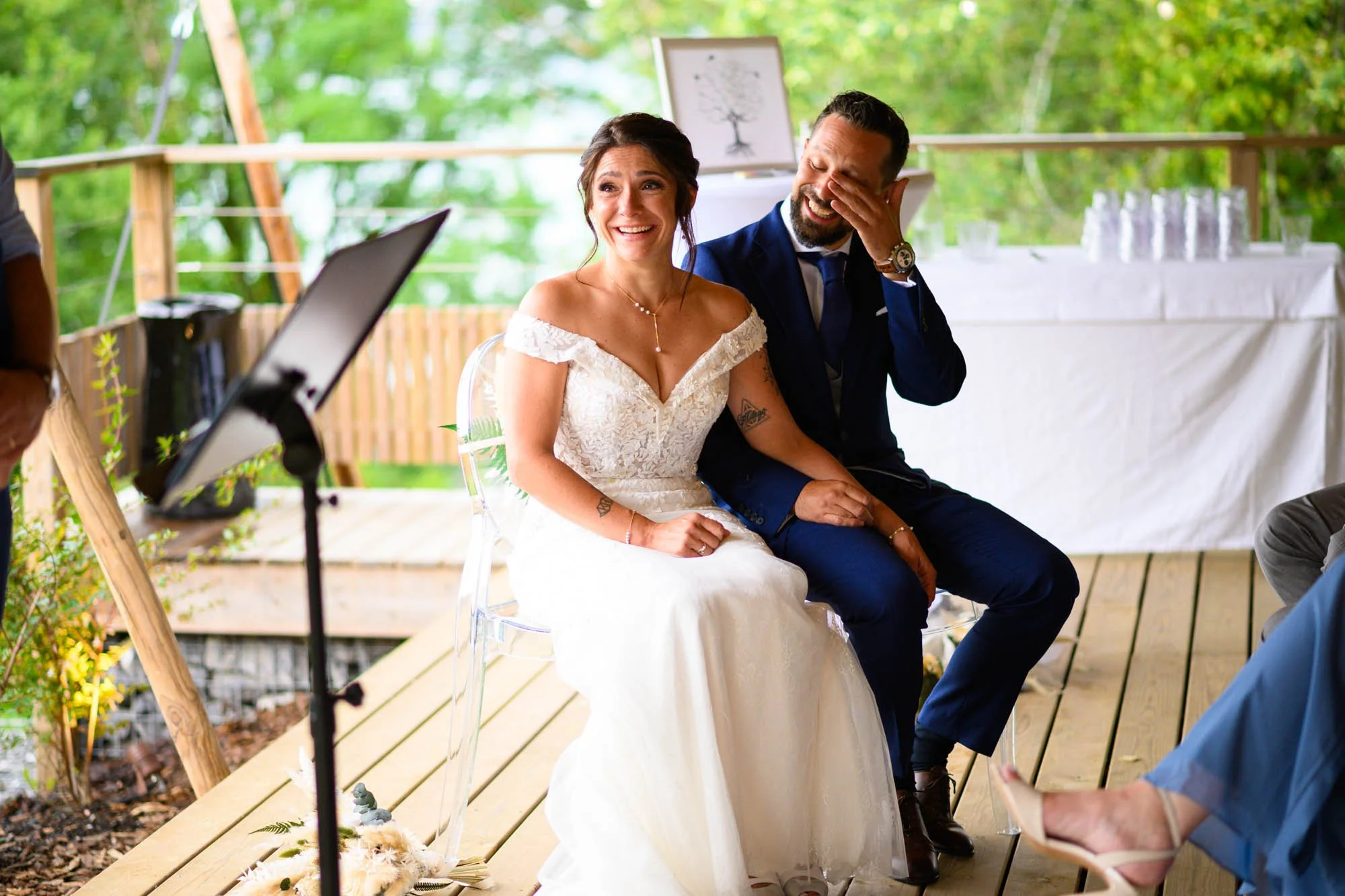 Un couple de mariés assis lors de leur mariage en plein air, la mariée en robe blanche et le marié en costume bleu, ils rient et partagent un moment de joie.