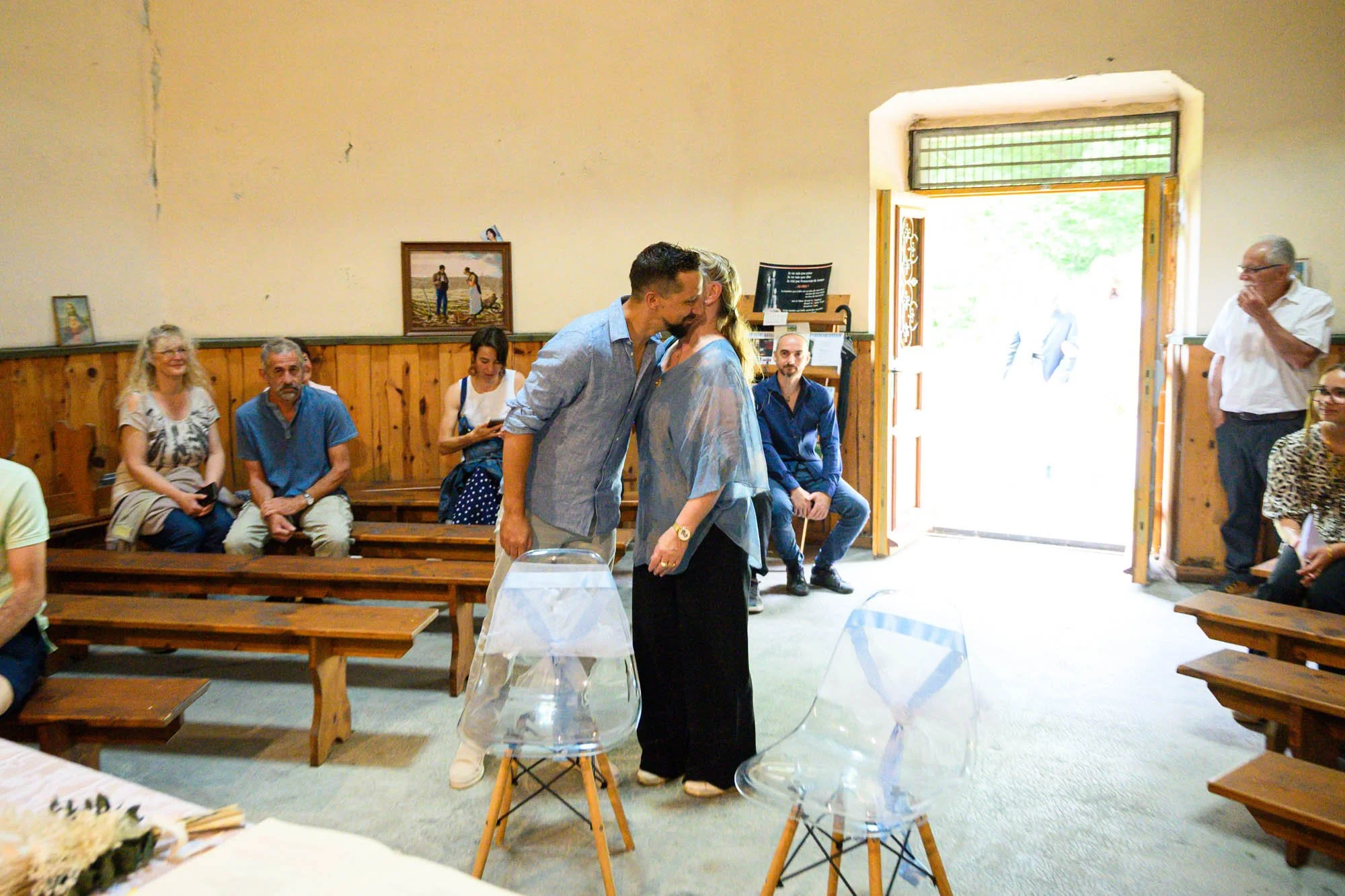 Un couple s'embrasse dans une salle où assiste à une cérémonie. Plusieurs personnes sont assises sur des bancs en bois, certaines regardent le couple, d'autres utilisent leur téléphone. La salle a des murs en bois et une porte ouverte laissant entrer