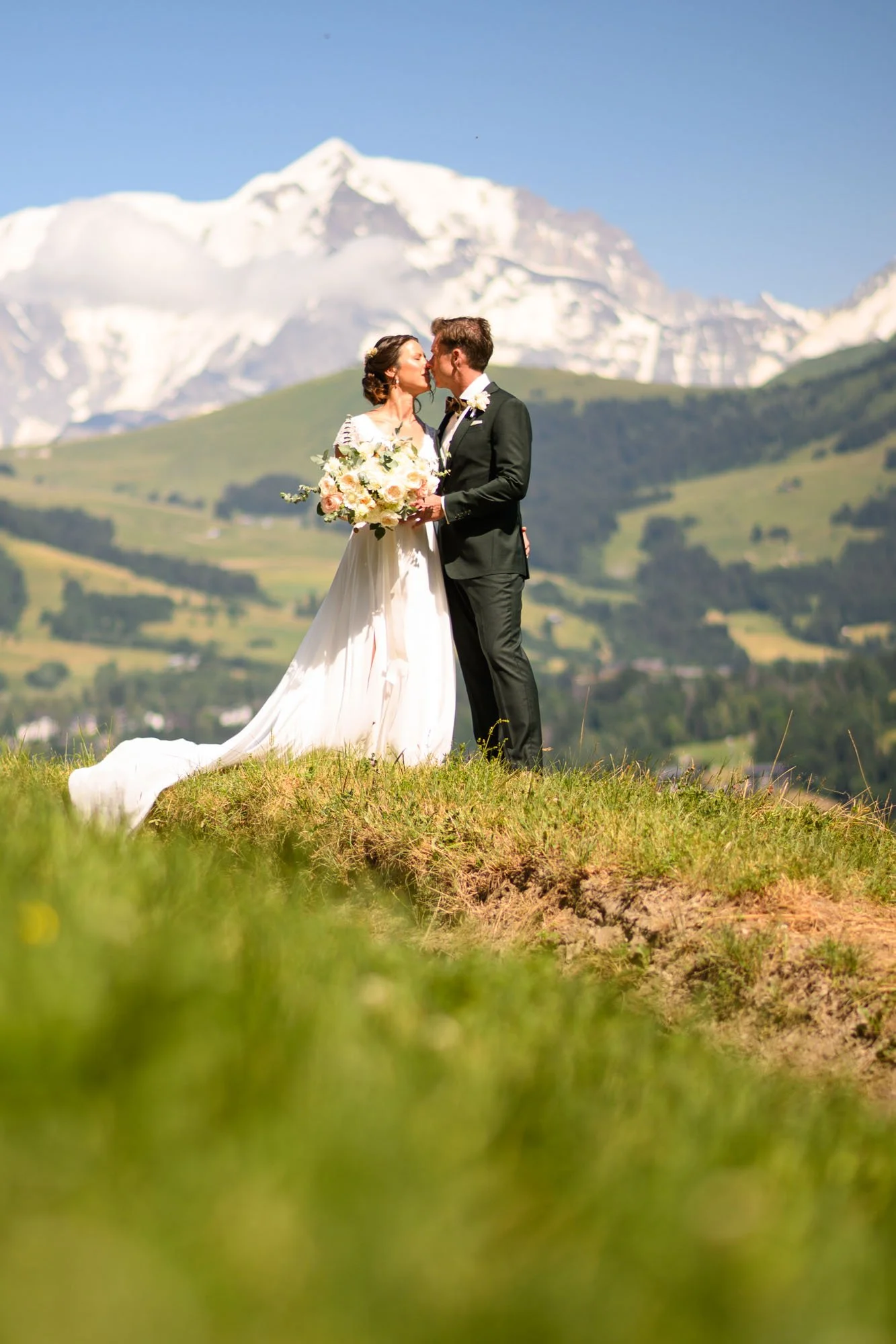 Un couple de mariés en costume et robe de mariée s'embrassant dans un paysage de montagnes et de collines verdoyantes.