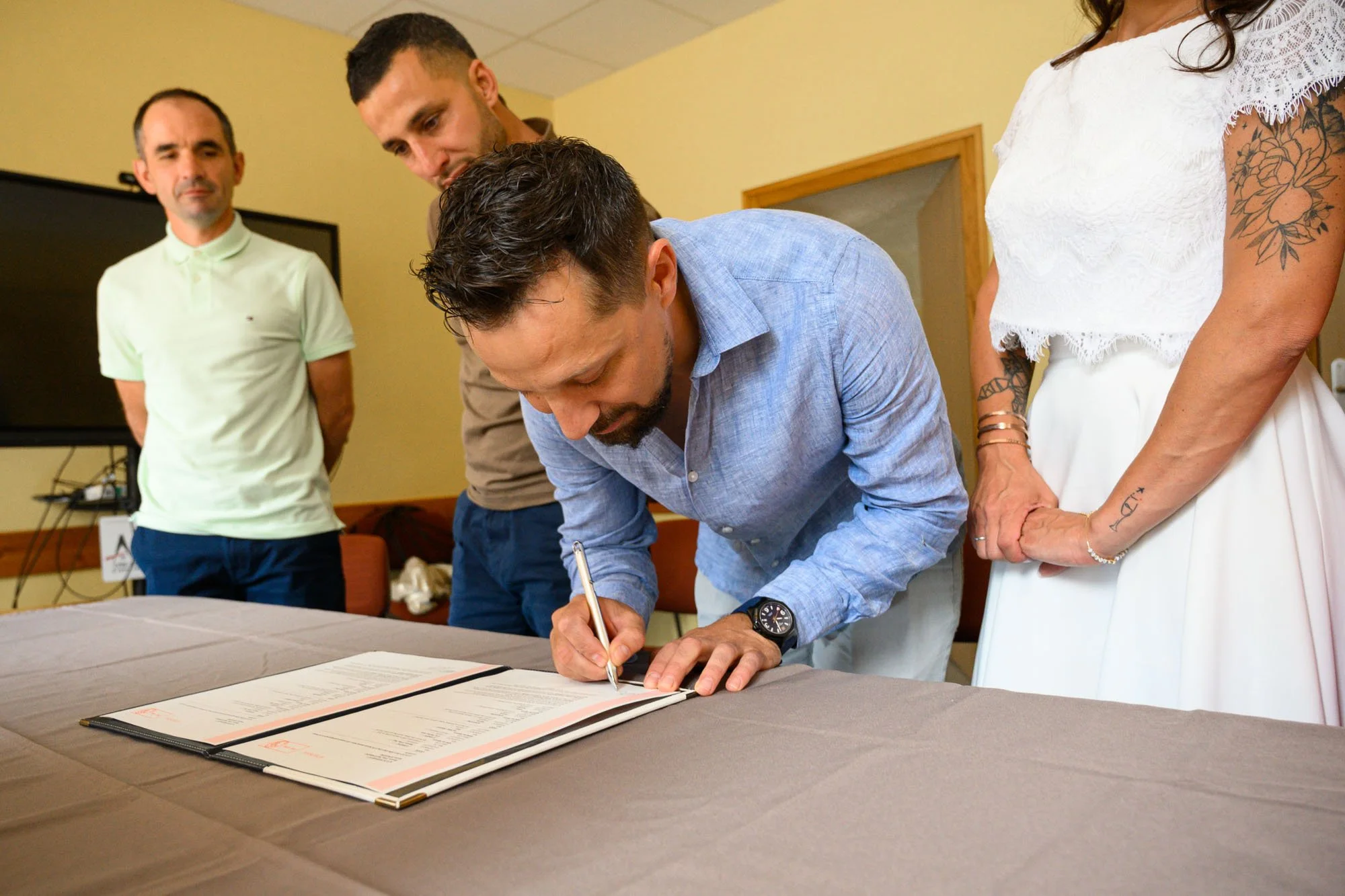 Un homme signe un document lors d'une cérémonie, entouré de trois autres personnes dans une salle intérieure.