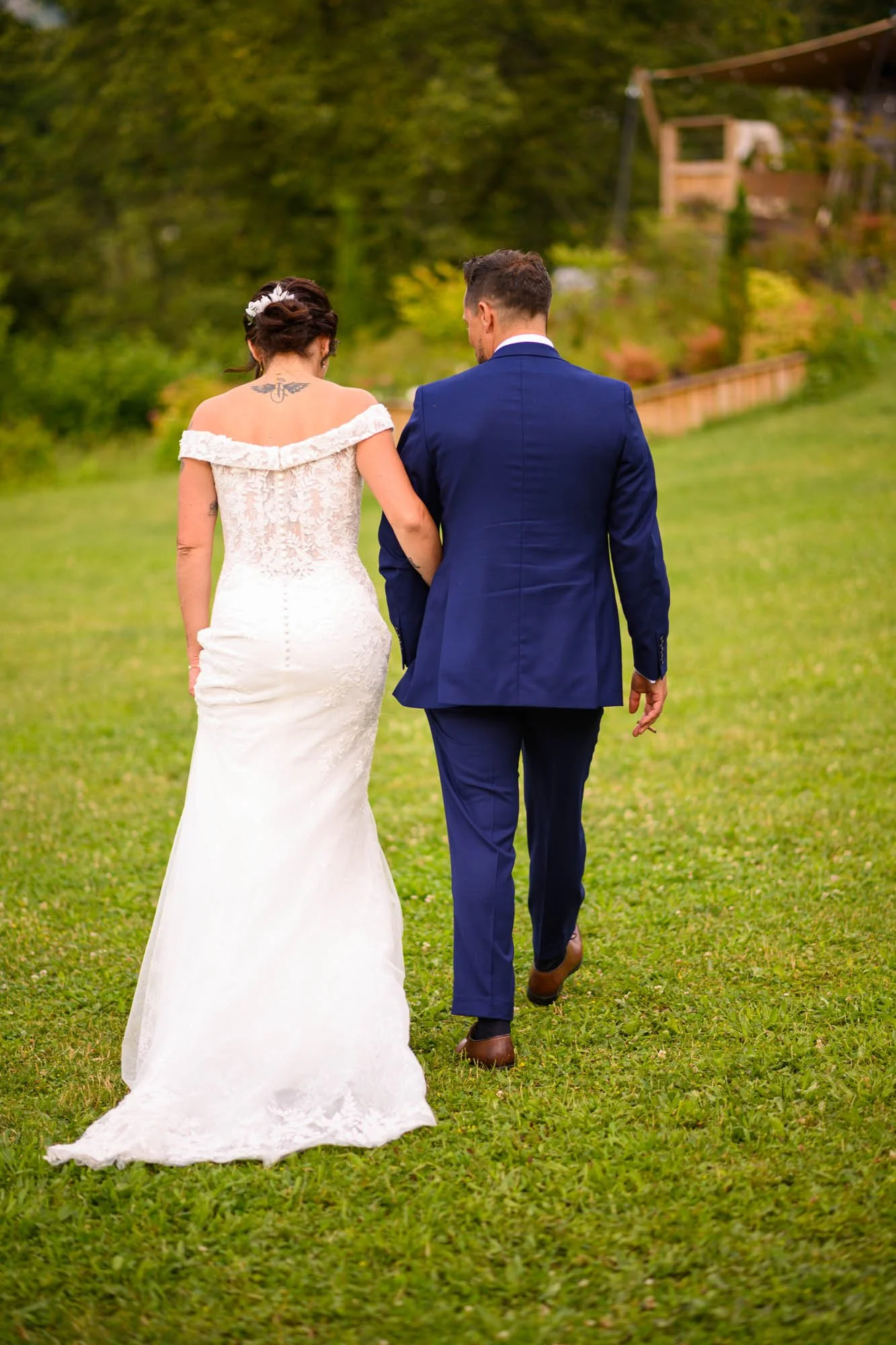 Un couple marié marche dans un jardin verdoyant, la femme porte une robe de mariage blanche et un tatouage dans le dos, l'homme porte un costume bleu.