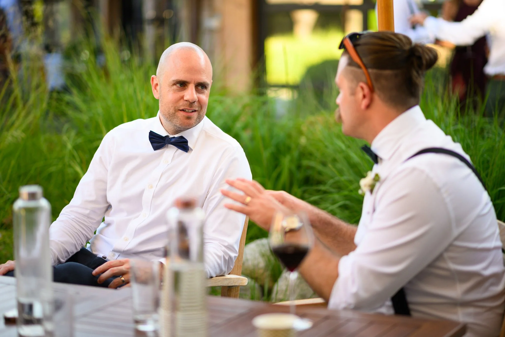 Deux hommes en tenue formelle, assis à une table en extérieur, discutent. L'un porte un noeud papillon, l'autre porte des lunettes de soleil sur la tête, avec une coupe de cheveux vintage, en train de parler.