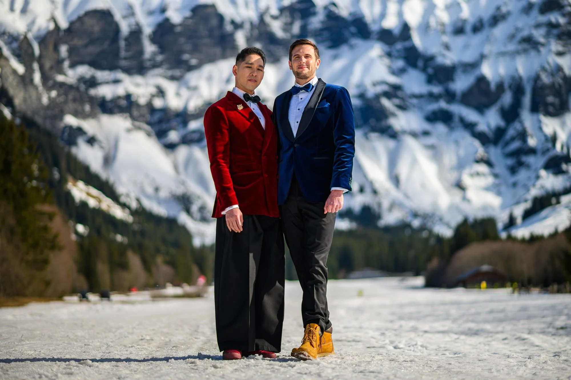 Deux hommes en costume élégants se tiennent côte à côte dans un paysage enneigé avec des montagnes et des forêts en arrière-plan. L'un porte un costume rouge avec un nœud papillon noir, l'autre porte un costume bleu avec une chemise blanche et un nœu