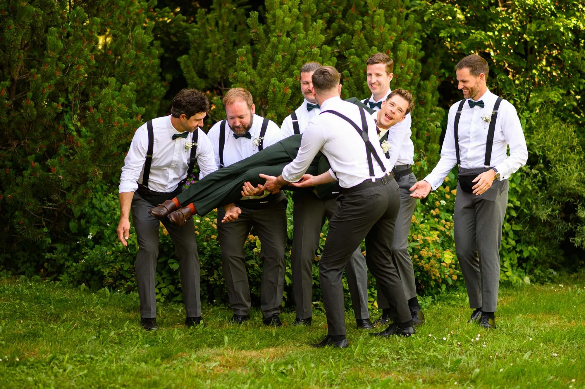 Groupe d'hommes habillés en costume et bretelles, jouant à un jeu d'amusement dans un jardin avec des arbustes verts en arrière-plan, lors d'une occasion festive.