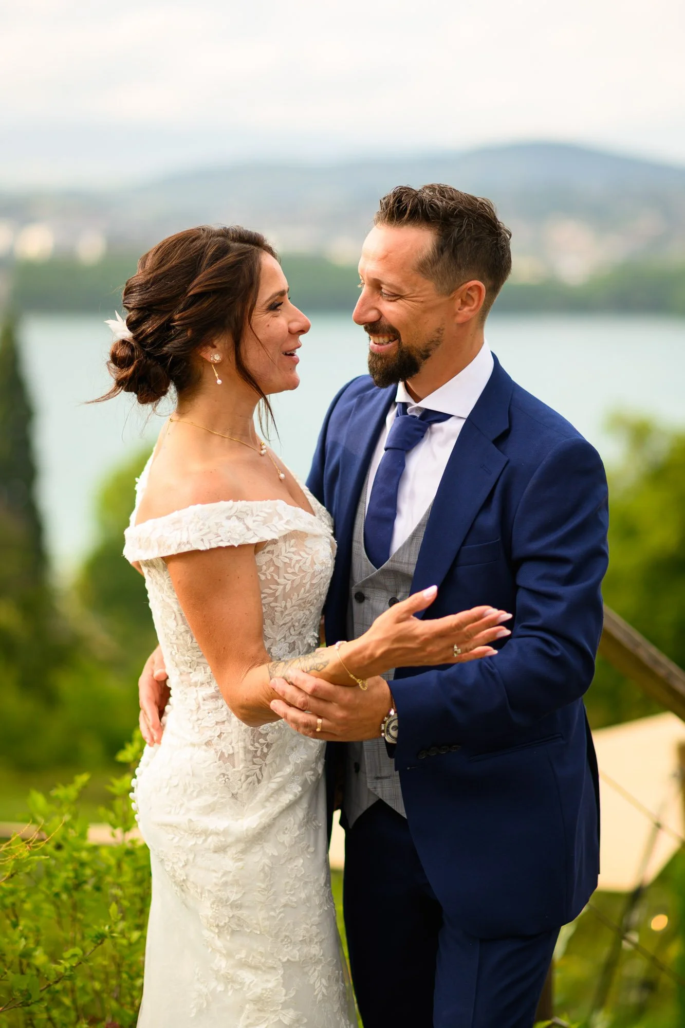 Un couple de mariés souriant et se tenant par la main à l'extérieur, avec un lac et une montagne en arrière-plan.