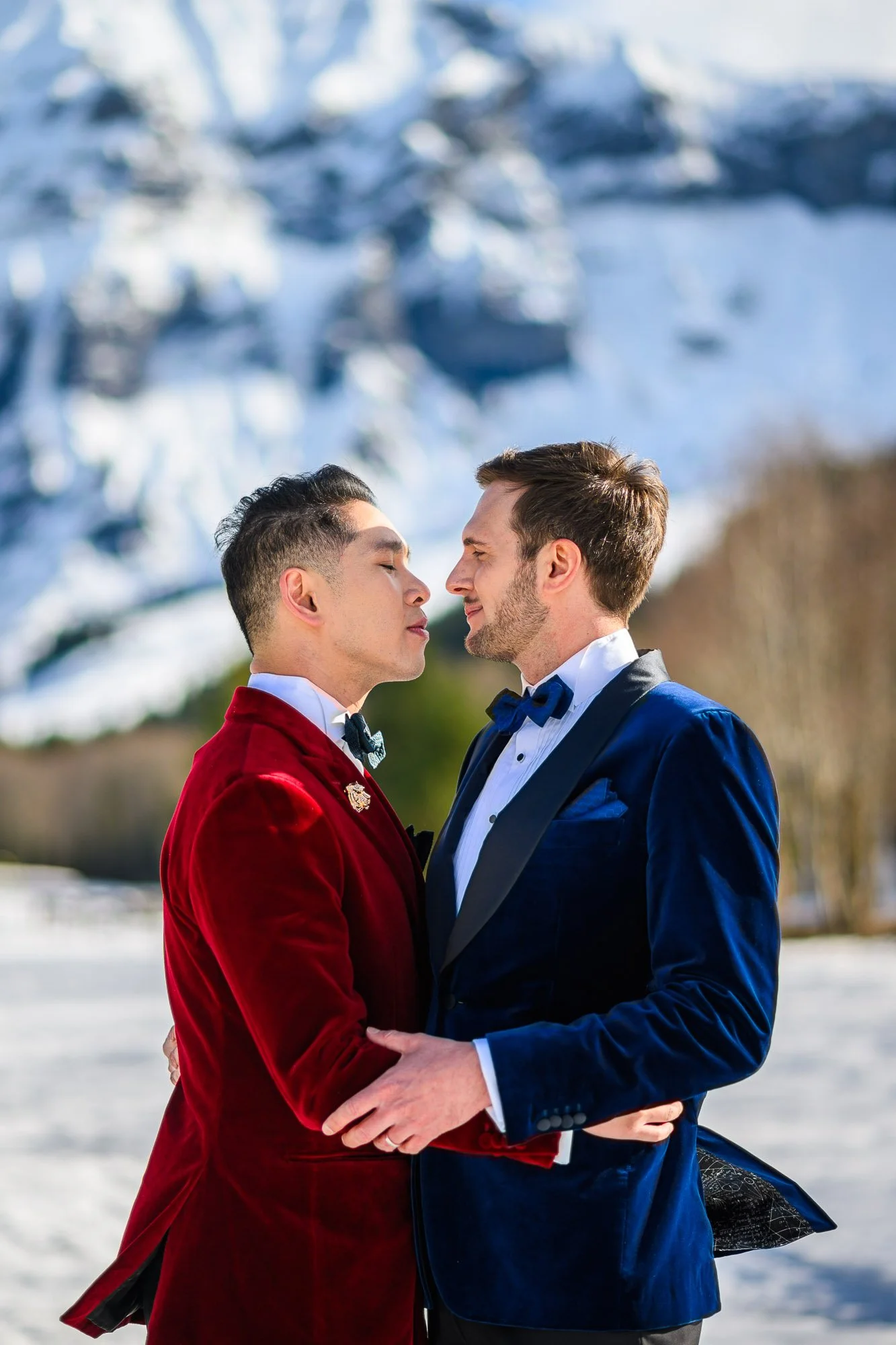 Deux hommes en smoking coloré s'embrassent tendrement en plein air, avec des montagnes enneigées en arrière-plan.