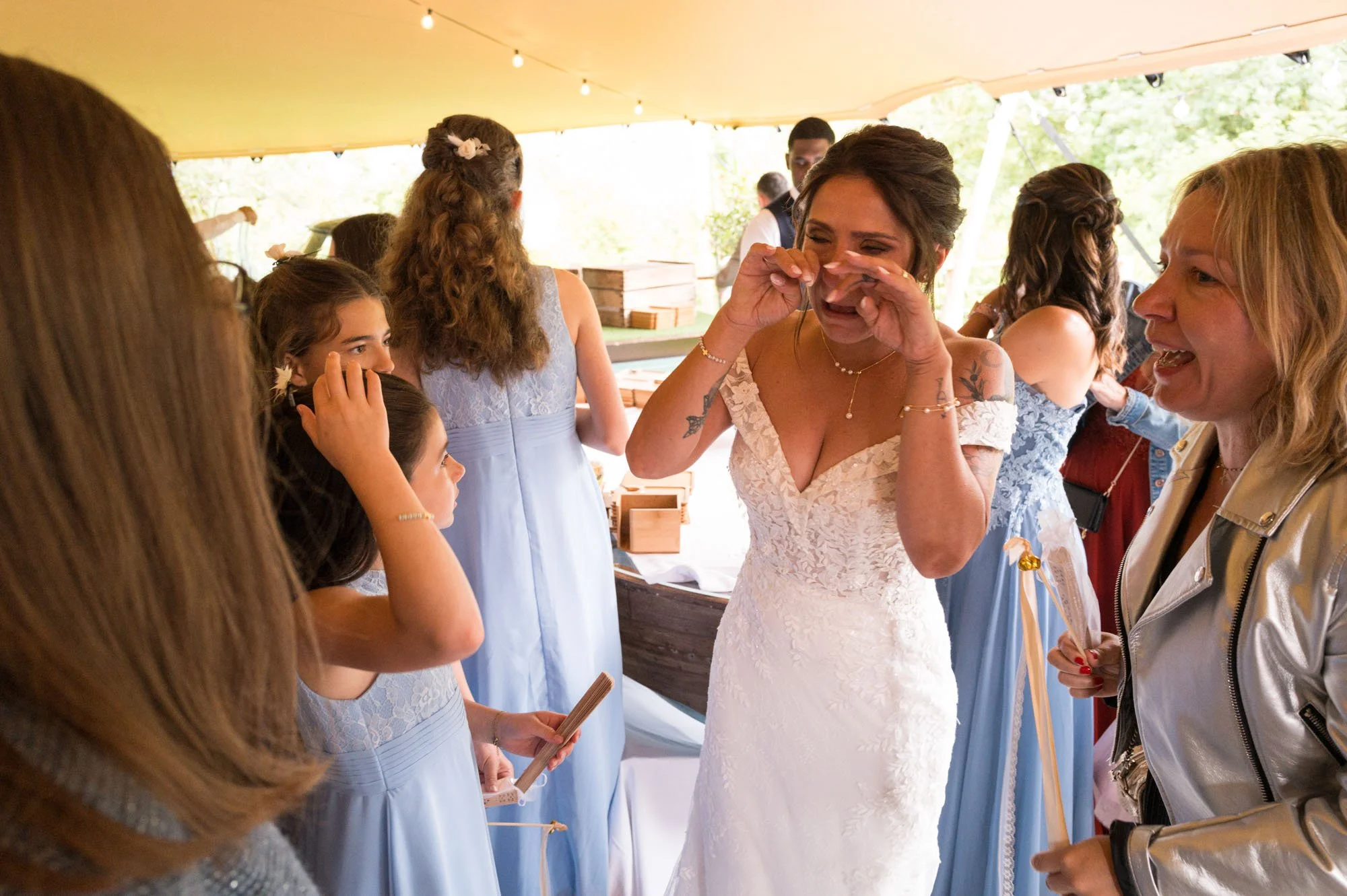Une femme en robe de mariée pleure d'émotion entourée d'autres femmes lors d'une fête en plein air sous une tente.