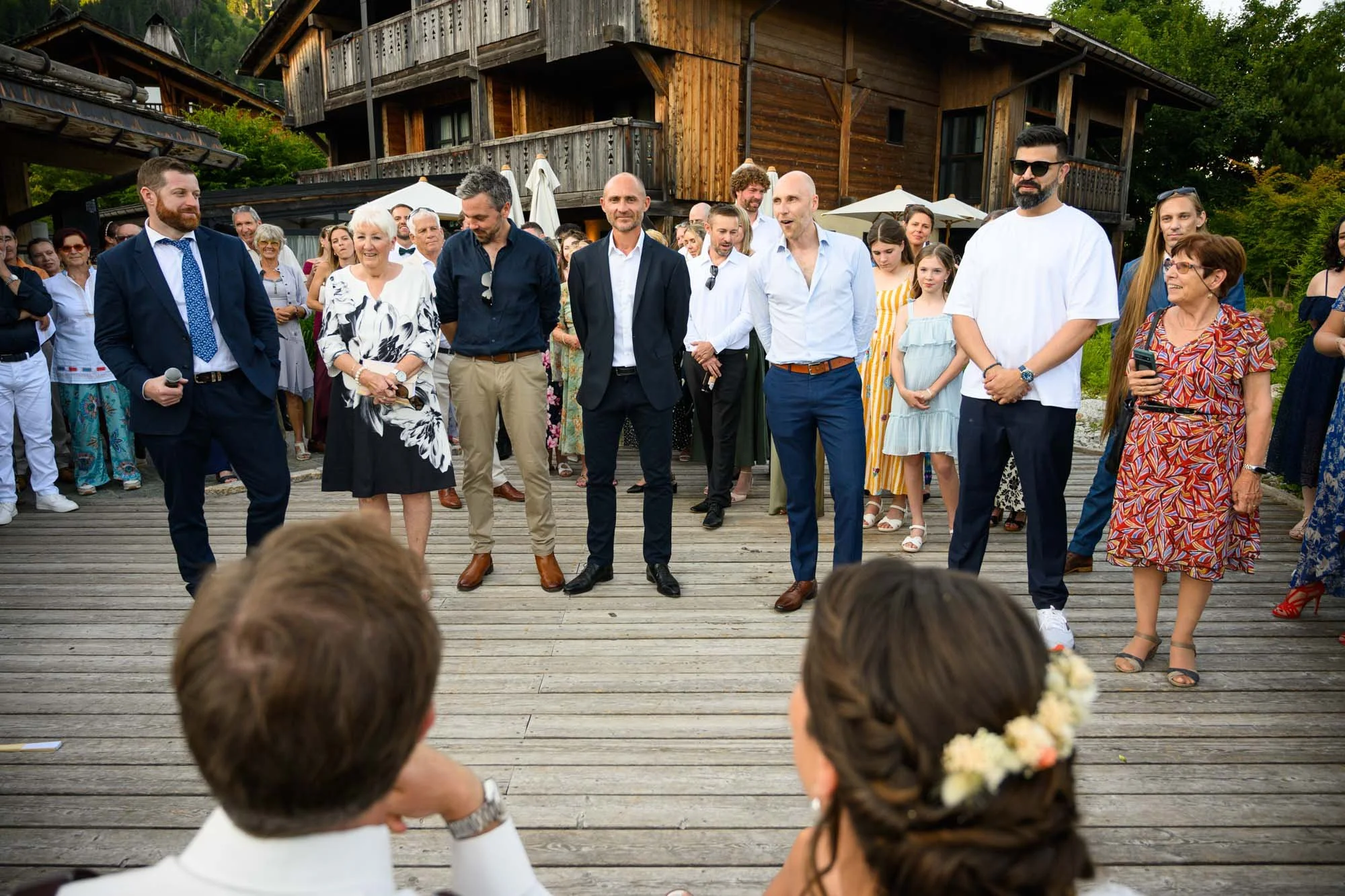 Groupe de personnes lors d'une célébration en plein air devant une maison en bois, certains dansent au centre, d'autres regardent et prennent des photos.