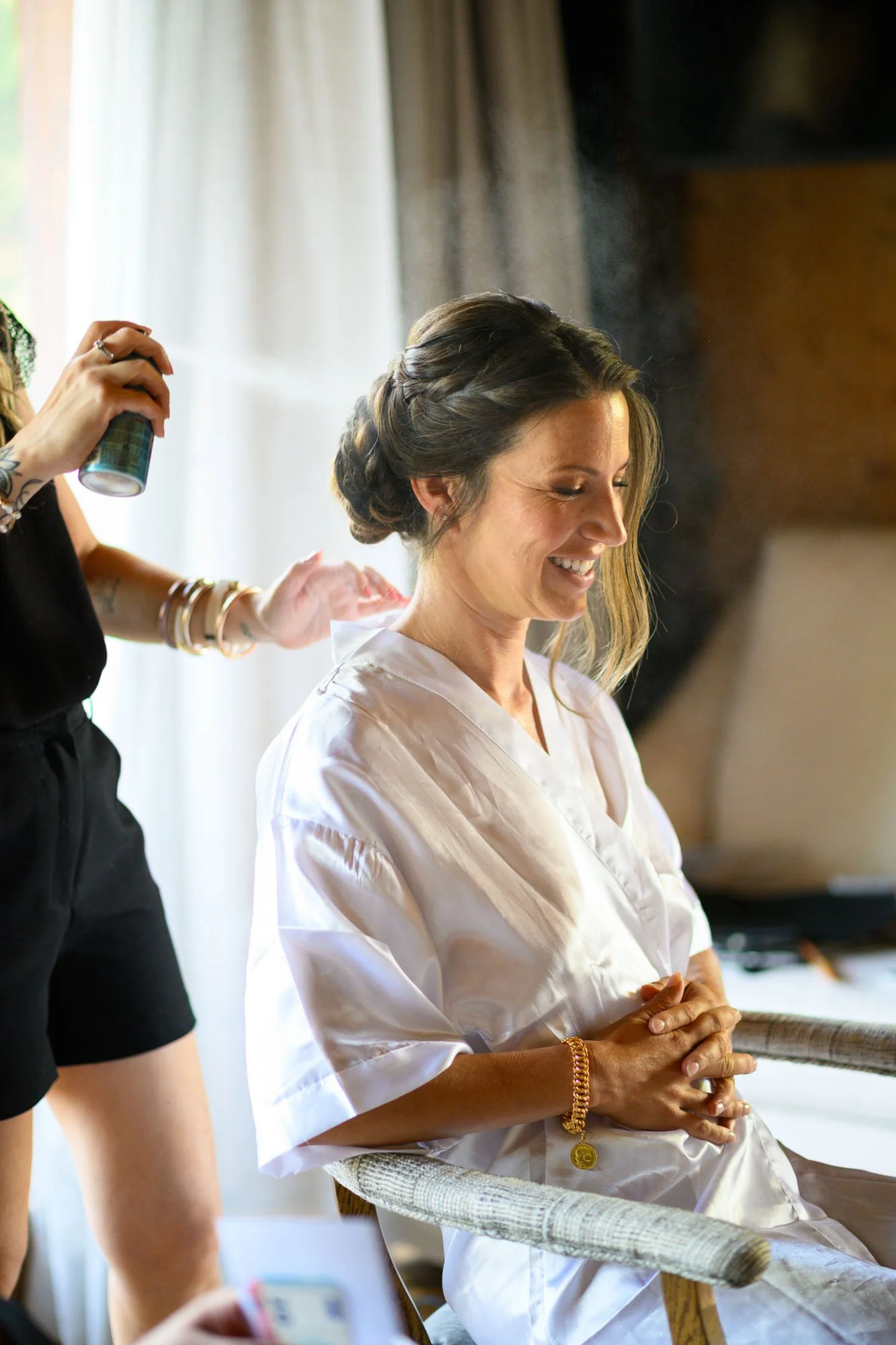 Une femme en robe blanche sourit en étant coiffée, une personne sprayant des produits de coiffure à côté, dans une pièce lumineuse.