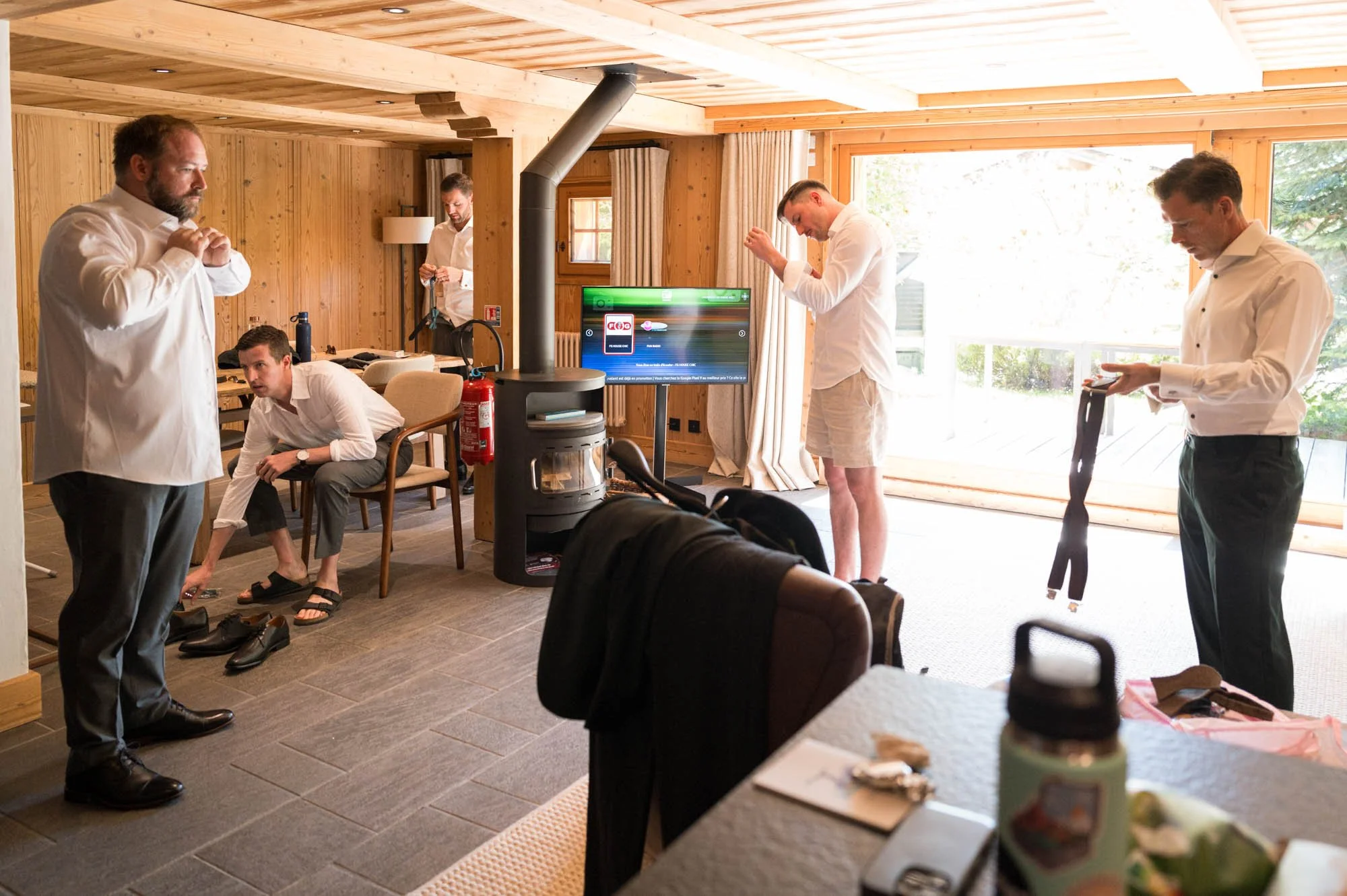 Groupe d'hommes en préparation dans une pièce en bois, certains ajustant leurs vêtements, d'autres utilisant leurs téléphones, avec un grand balcon lumineux derrière.