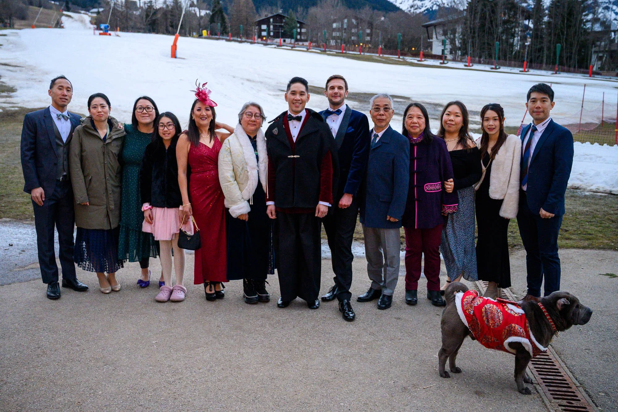 Groupe de personnes posant pour une photo devant une zone enneigée, avec un chien portant un manteau rouge à motifs, lors d'un événement familial ou festif.